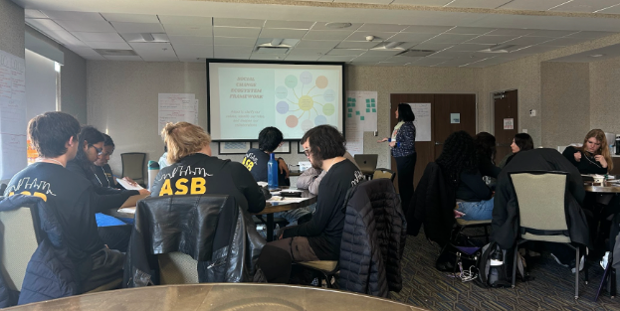 A group of students sitting at tables, focused on material in front of them, while Deepa Iyer stands among them as if leading a class