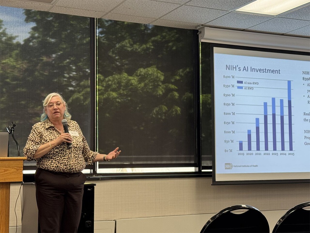 Dr. Susan Gregurick stands beside a podium in front of a digital display with a bar graph visualization with title, "NIH's AI Investment."