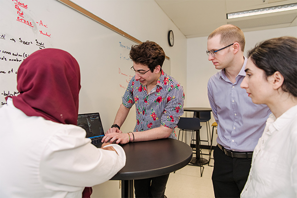 Group in discussion gathers around computers. Equations on whiteboard in background.