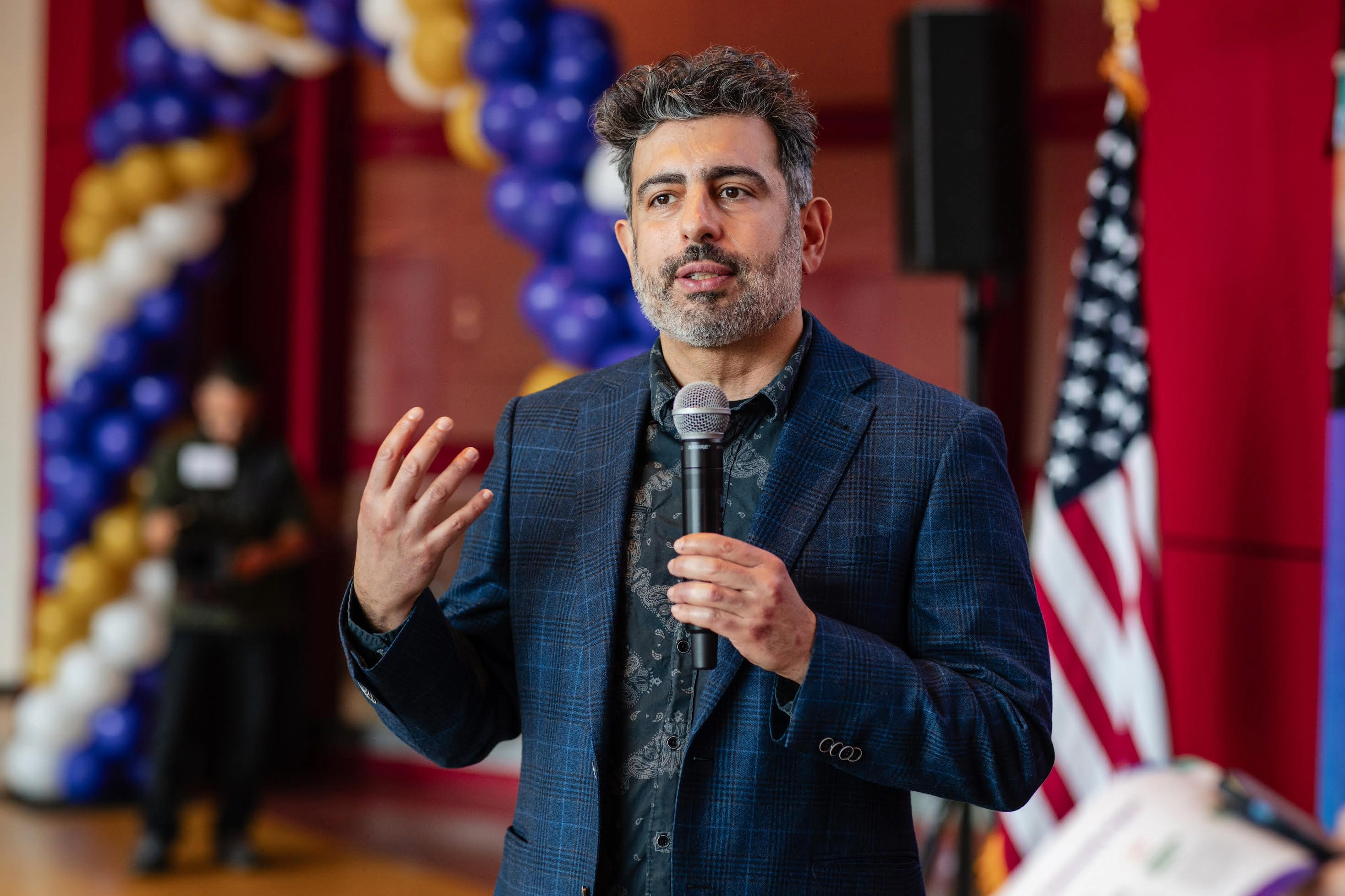 A man in a plaid blazer speaks into a microphone indoors, with U.S. flags and balloon decorations in the background.