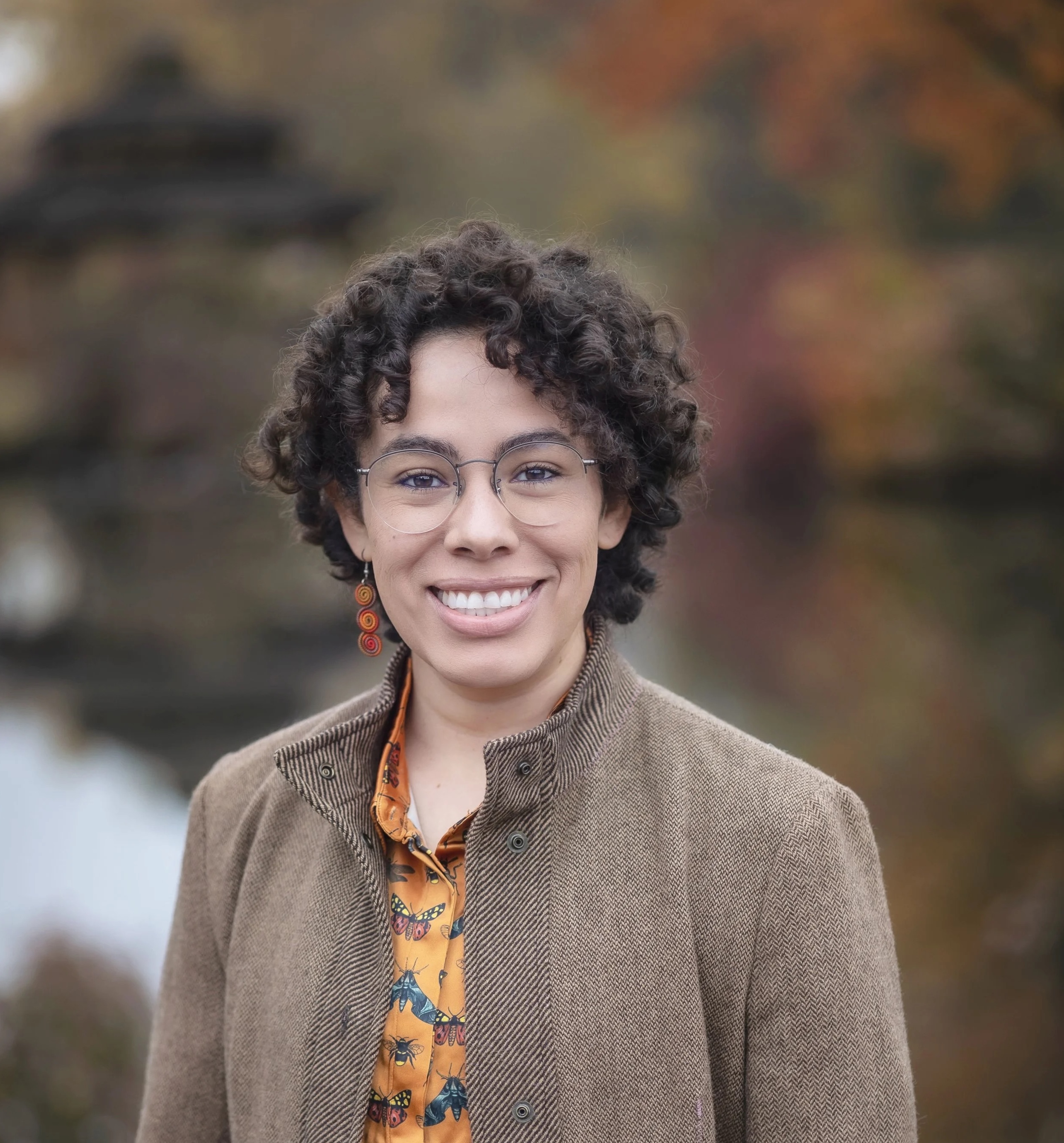 A Latine person with brown curly hair is smiling. They are wearing glasses and a dangly earring in their right ear.