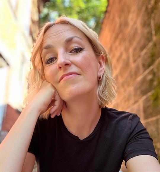 A white woman with short blond hair rests her face on her hand and looks forward in a downwardly direction.