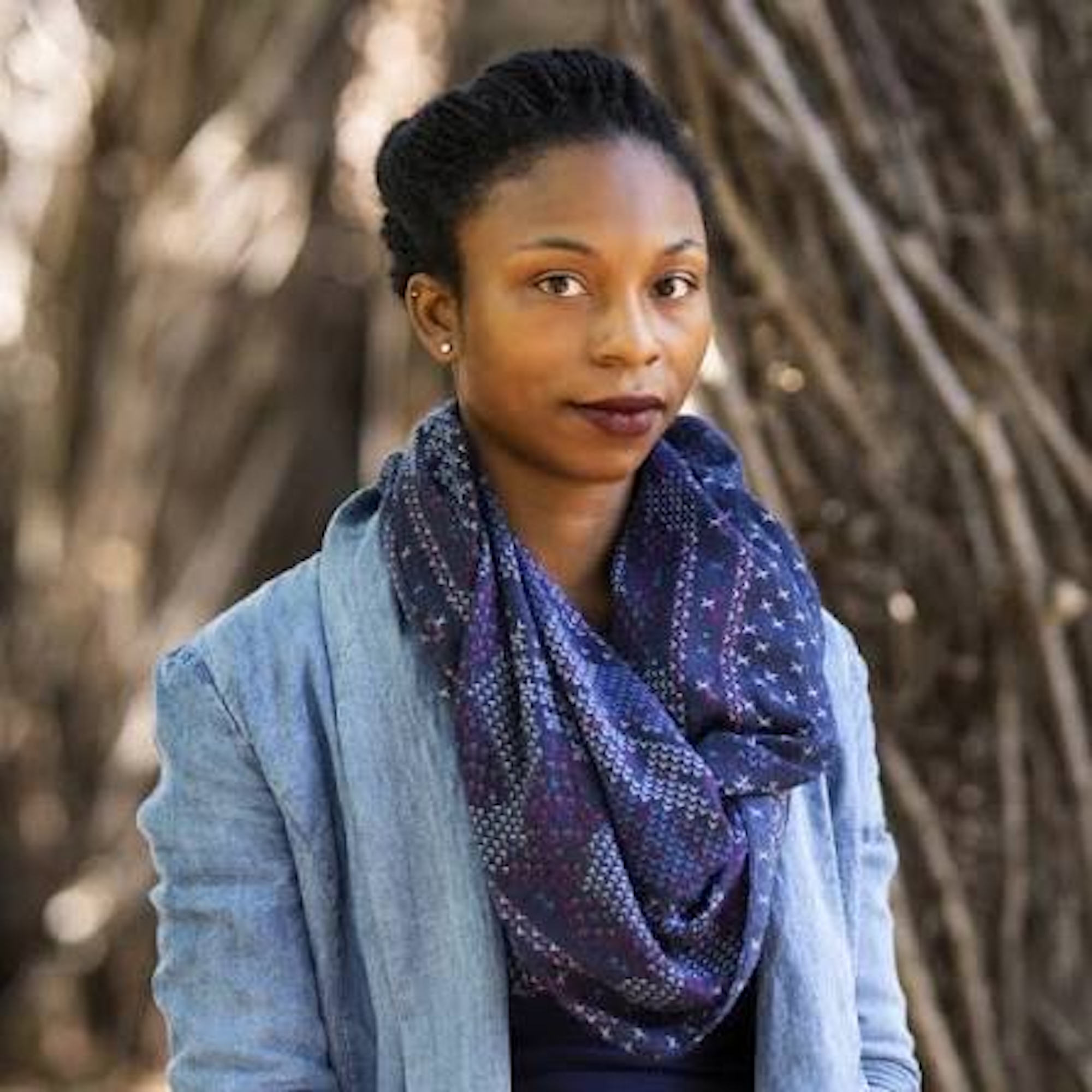 A Black woman wearing a blue blazer and a big blue scarf is looking at the camera.