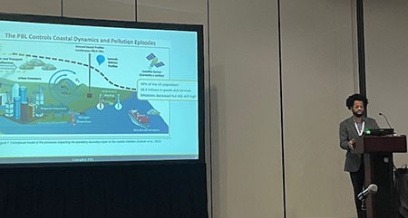 Maurice Roots stands at a podium and presents his research, which is on a screen to his right.