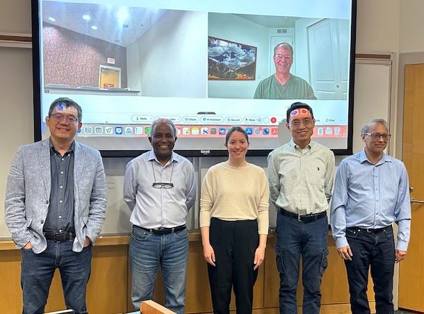 Drs. Zhang, Demoz, Zhai, Desouza-Machado standing in front of a screen, all wearing long sleeve button downs and jeans, with Kylie Hoffman in the center, wearing a pale yellow sweater, and Dr. Allen in a light green t-shirt, attending virtually