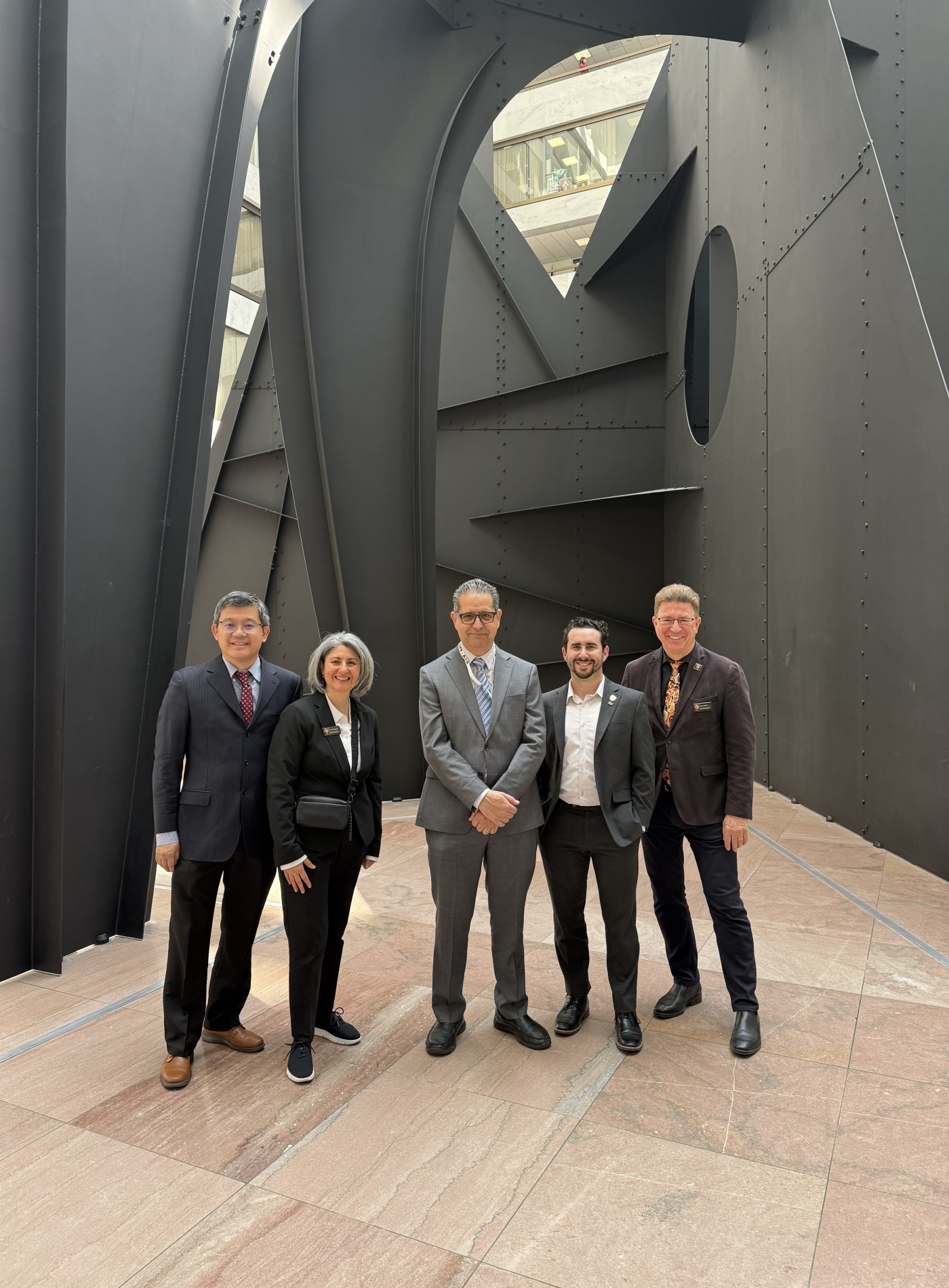 Five people in suits stand below a dark grey sculpture in an atrium.