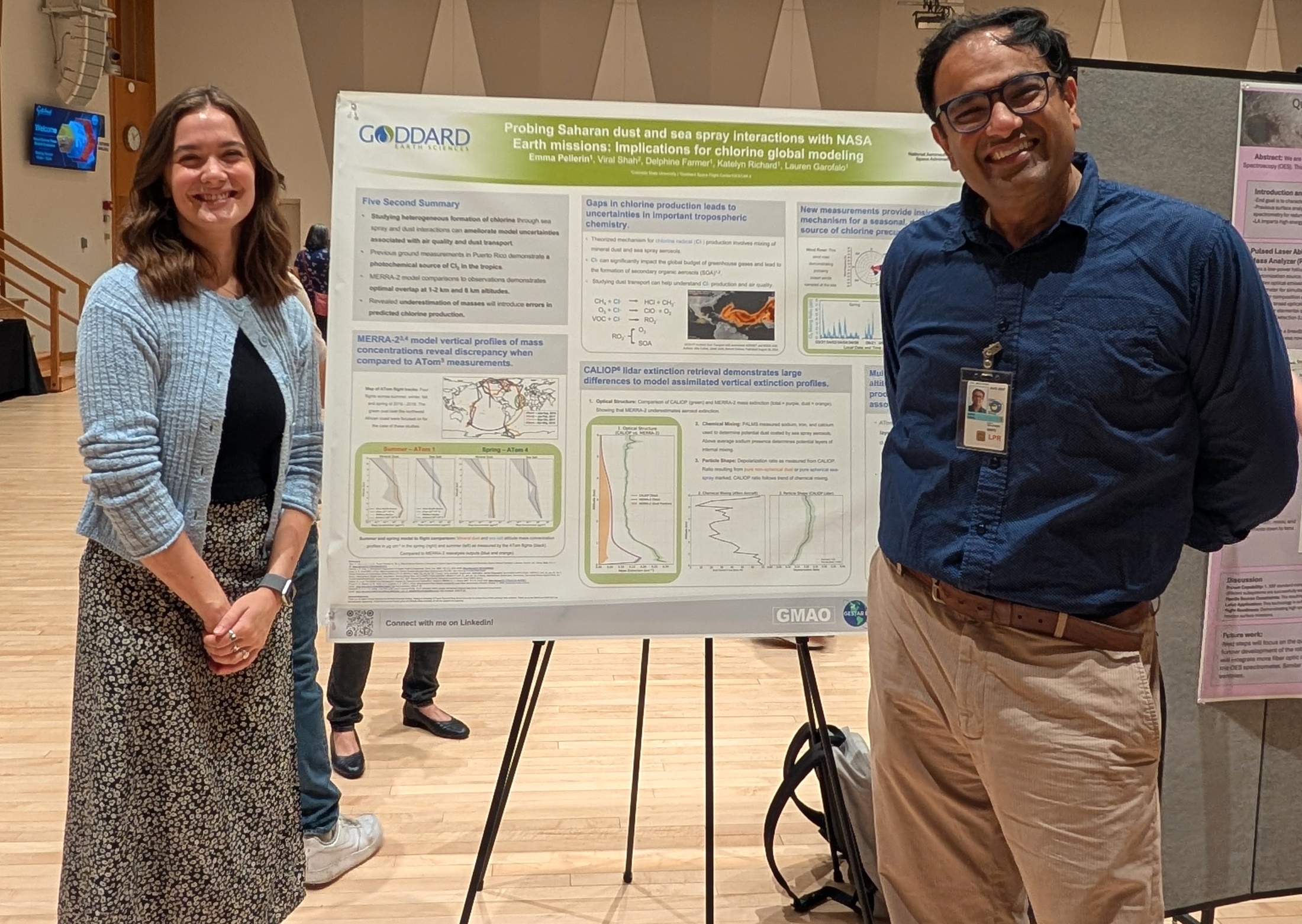 Emma in black tshirt and floral skirt along with Viral (Ver-all) Shah, in a button down and khakis, stand on either side of their poster in an auditorium with pale wood flooring