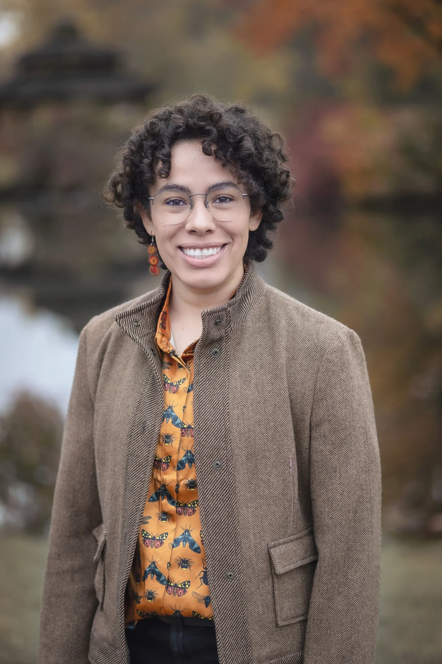 A Latine person with curly brown hair is smiling. They are wearing glasses and a brown jacket.