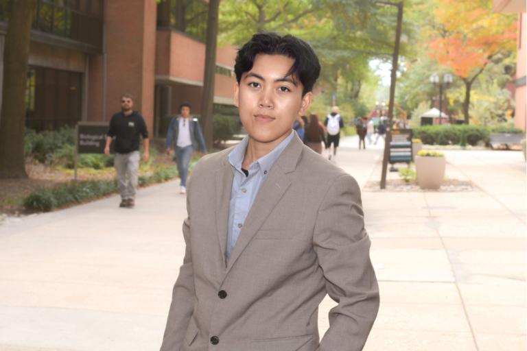 Thalia, wearing a tan blazer and a light blue button-down shirt, standing on the UMBC campus walkway. In the soft-focus background, other students are walking by.