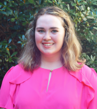Nicole wearing a bright pinks shirt smiling while standing in front of trees