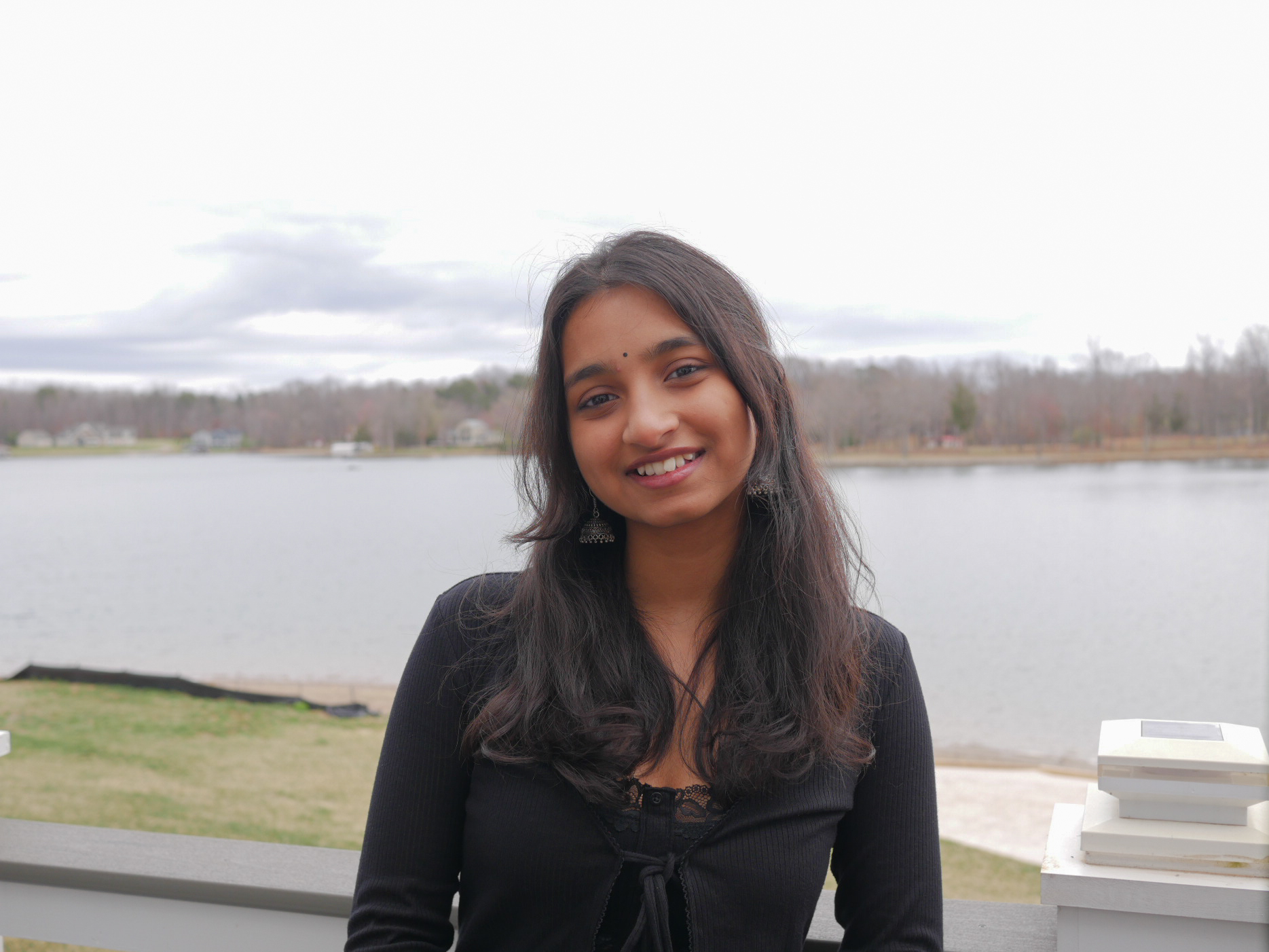 Trisha Tomcy smiling, standing outdoors with a lake and trees in the background under an overcast sky.