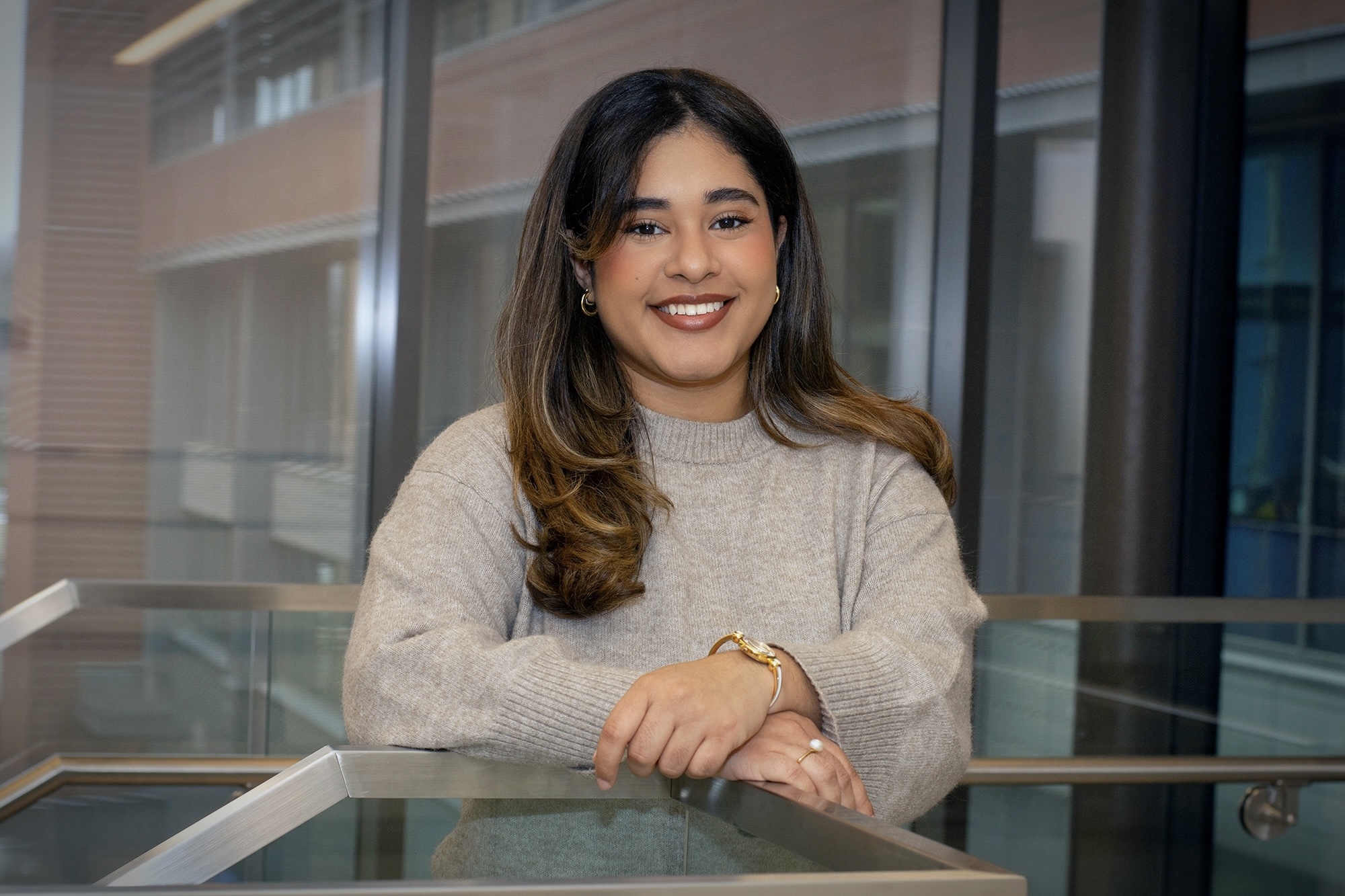 Michelle wearing a beige sweater, smiling and leaning against a glass railing in a campus building