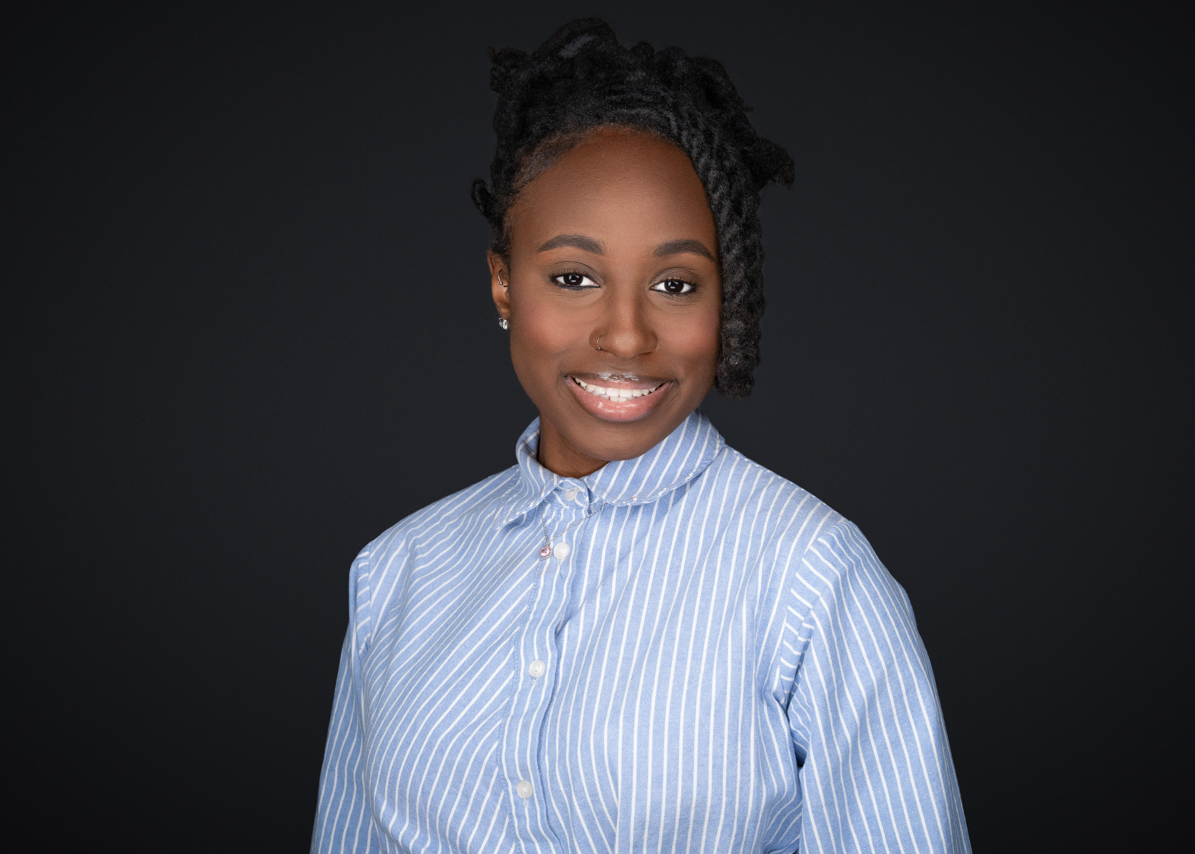 Aissatou smiling in front of a black background and is wearing a blue and white striped button up
