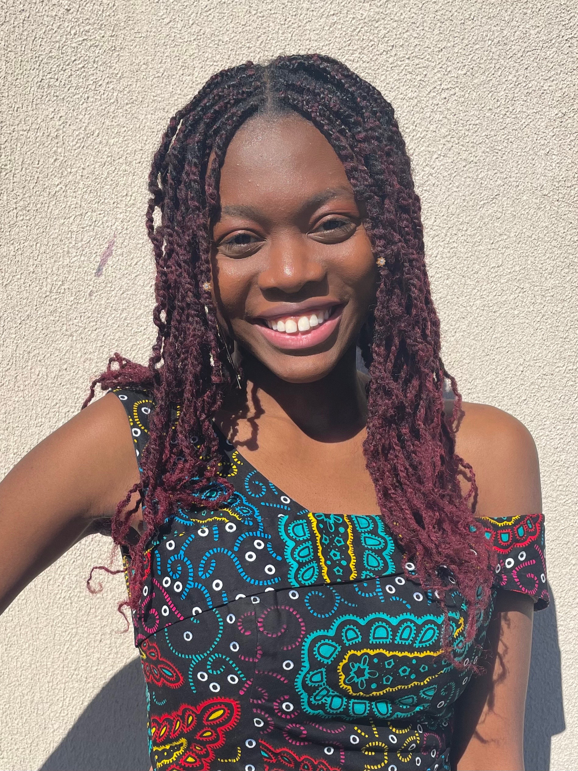 Joanna smiling against a light-colored wall. She is wearing a colorful patterned top with blue, yellow, and red paisley designs