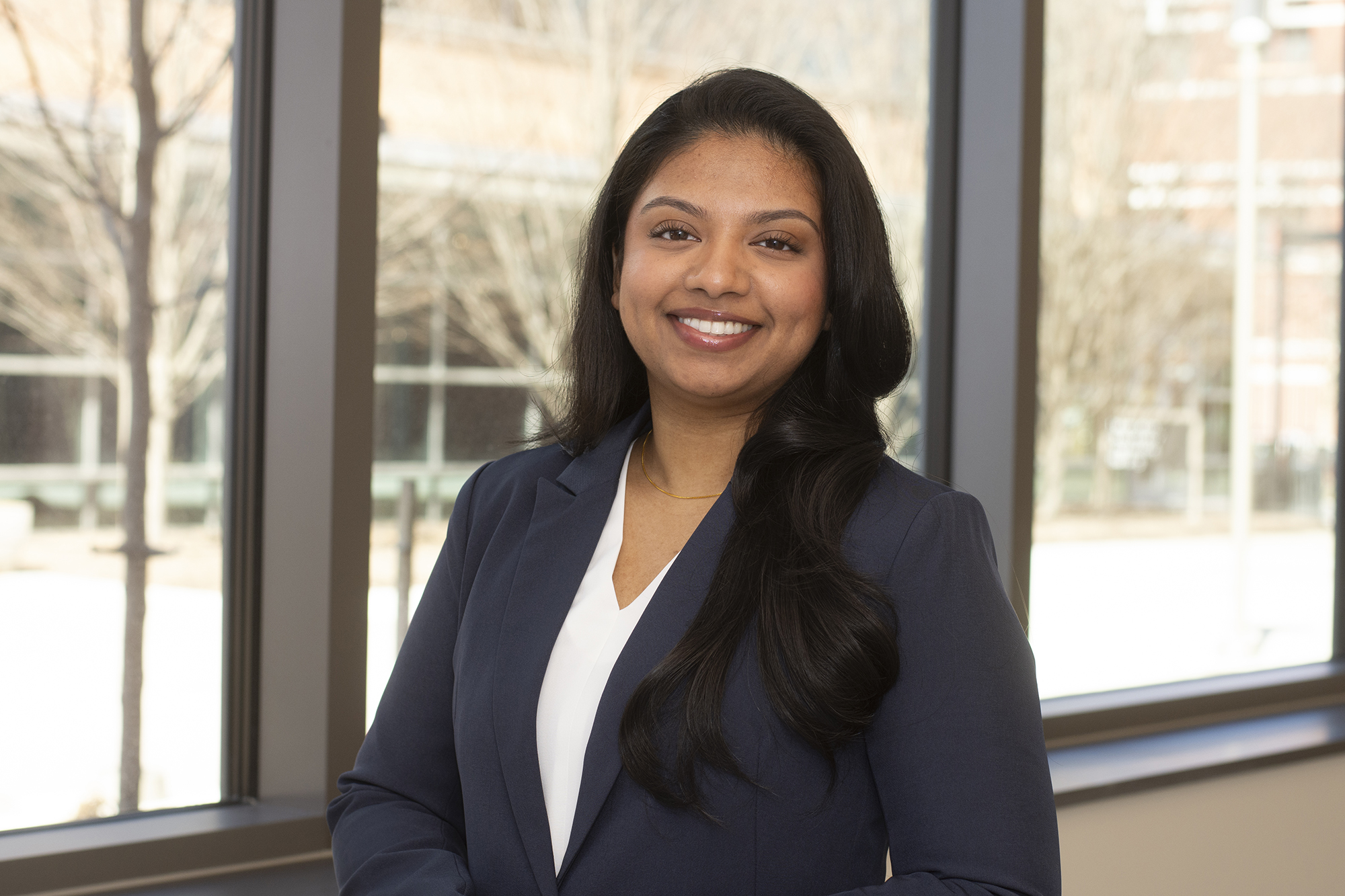 Noreen in a blue blazer with a white top smiling standing in front of glass window, with trees and a building outside