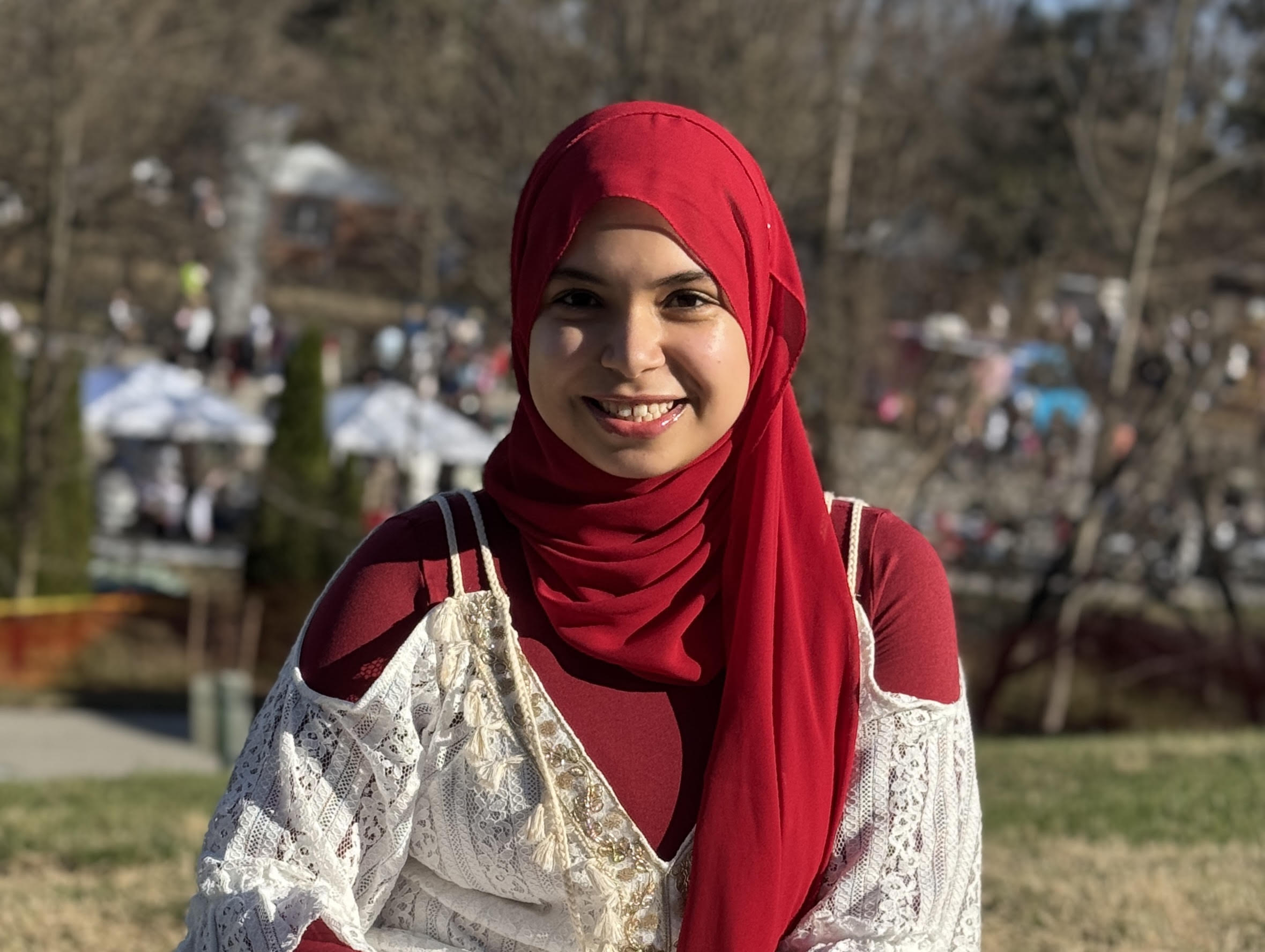 Aya in a red top and head scarf, smiling with people, tress and outdoor umbrellas blurred in the background