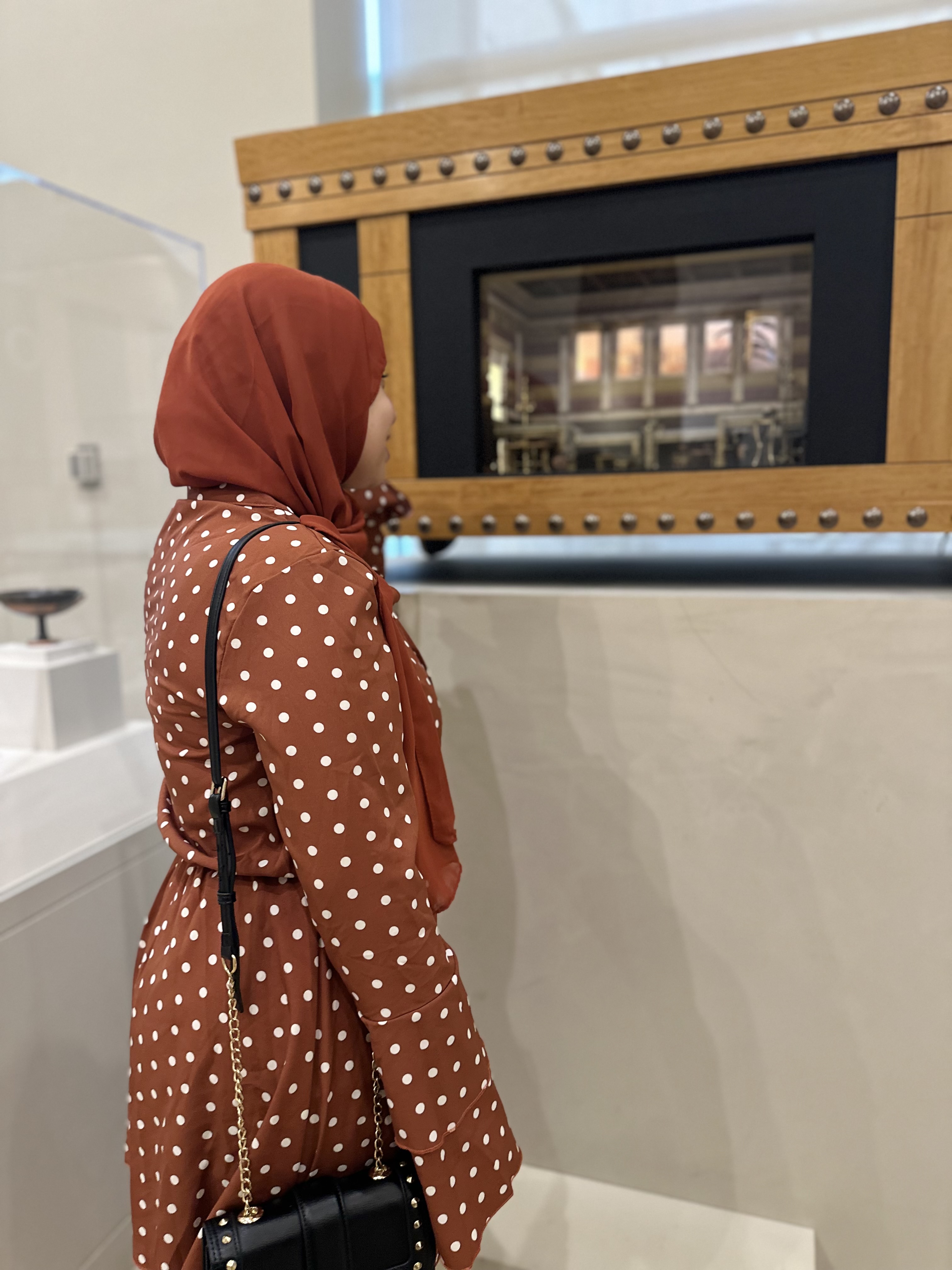 Aya in a polka dot dress and small black purse looking at A detailed miniature room diorama housed in a decorative wooden chest with metal rivets.
