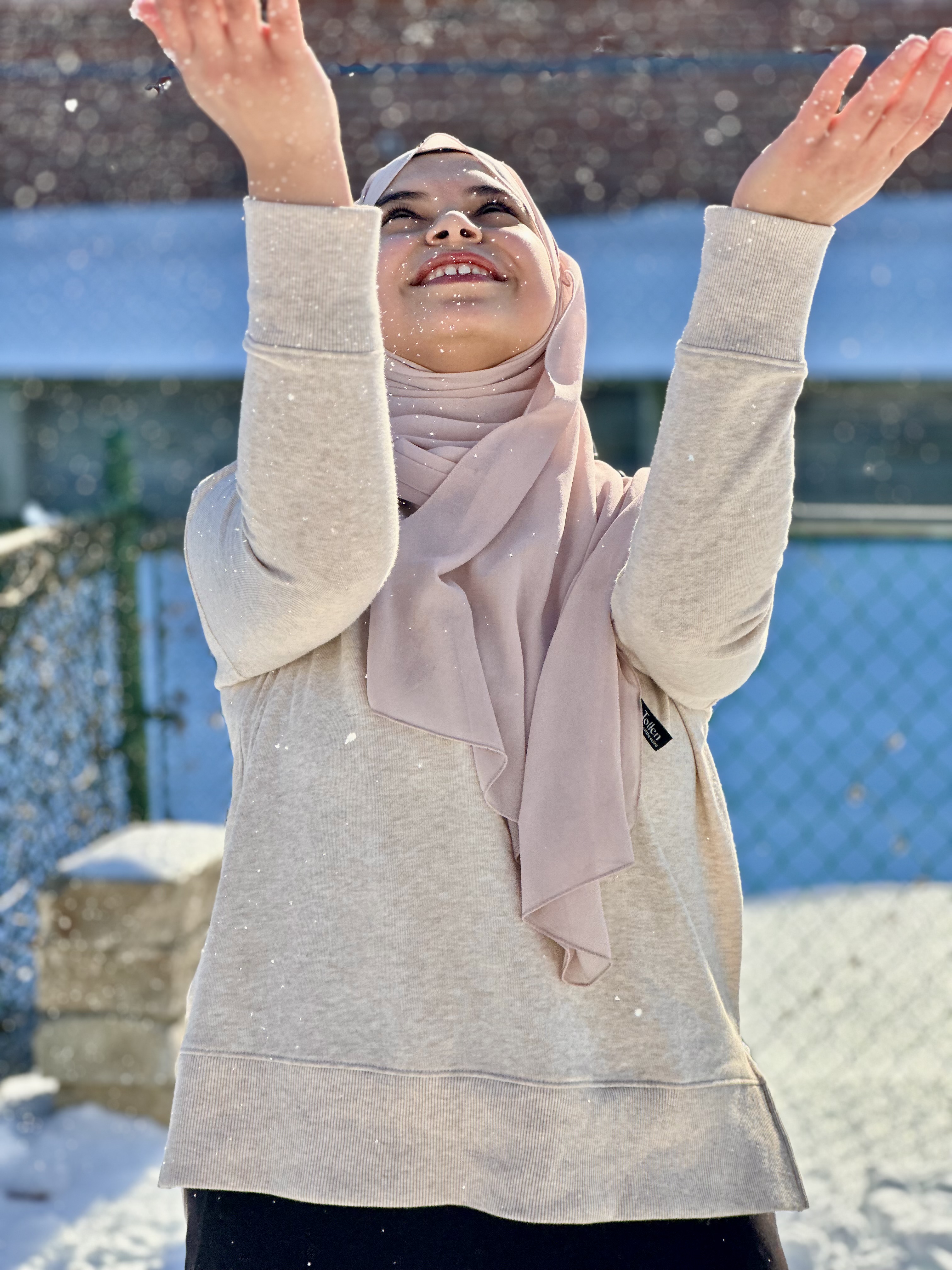 Aya standing outside looking up in the sky at the snow falling with hands up like, trying to catch the snow in the palm of their hands