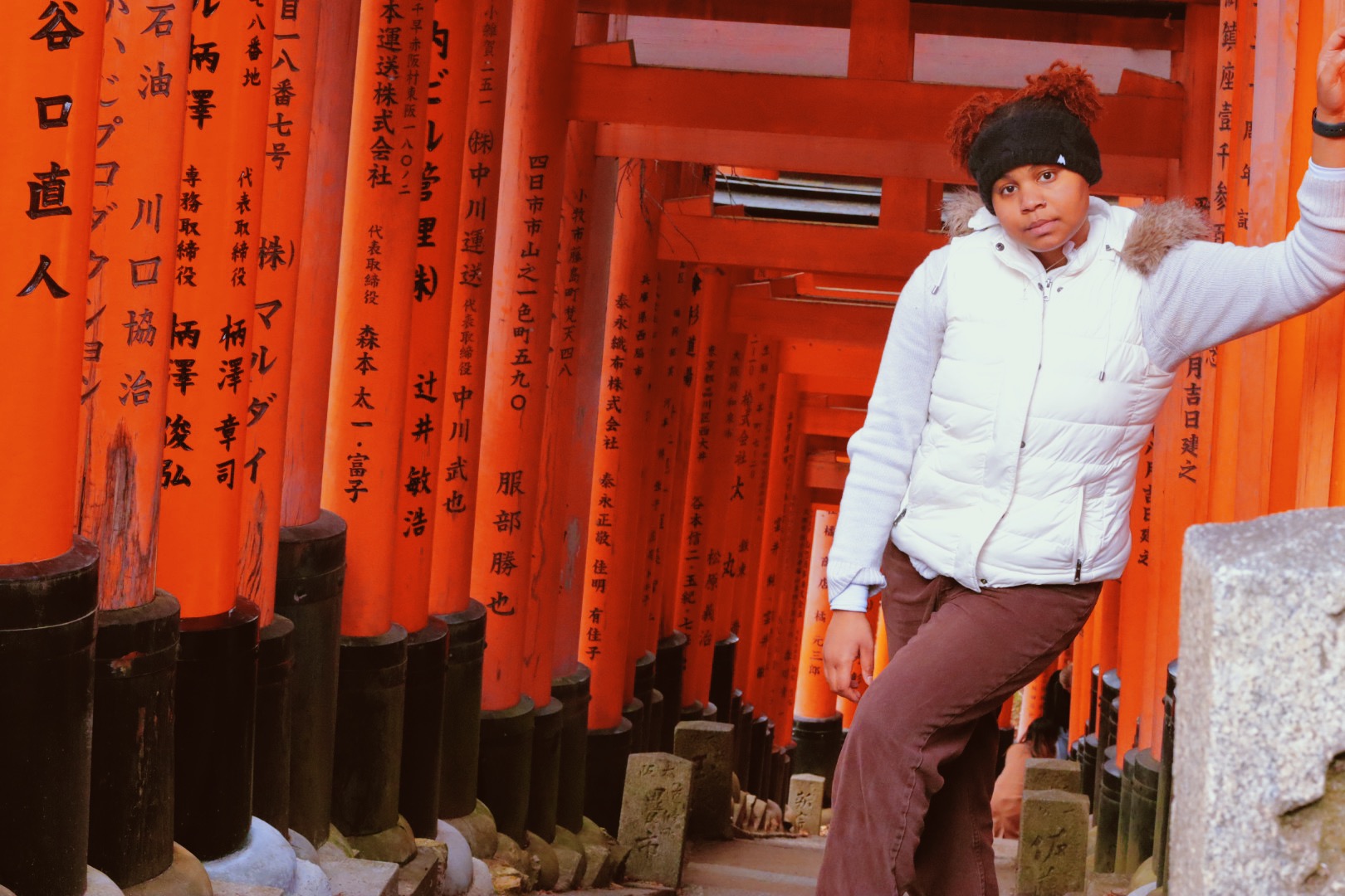 Shania in a whit coat and black hat with two pom poms, leaning on pole surrounded by a multiple red poles with character writing