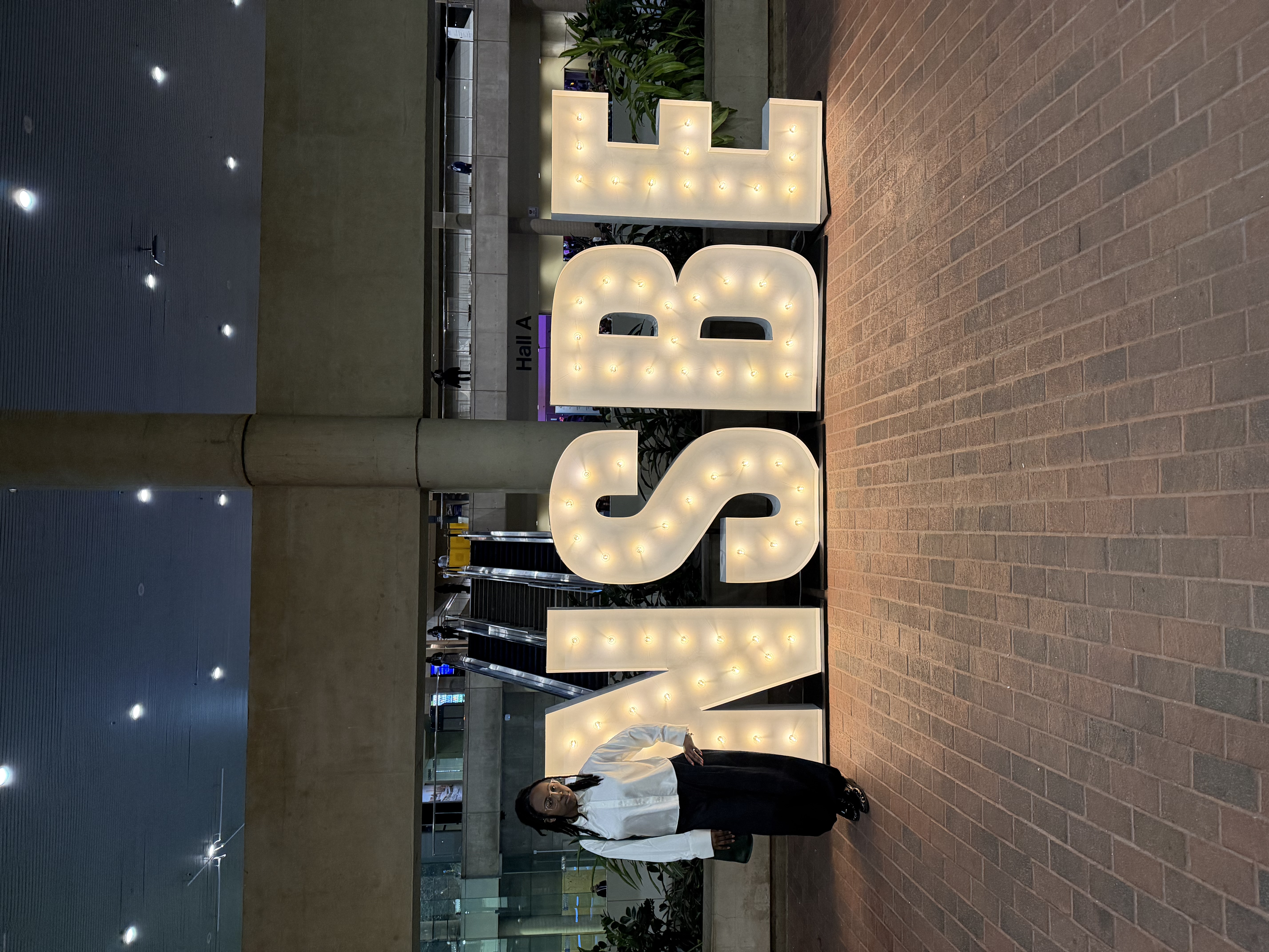 Aissatou in a button up collared shirt, smiling standing next to the big NSBE letters that have lights inside the letters