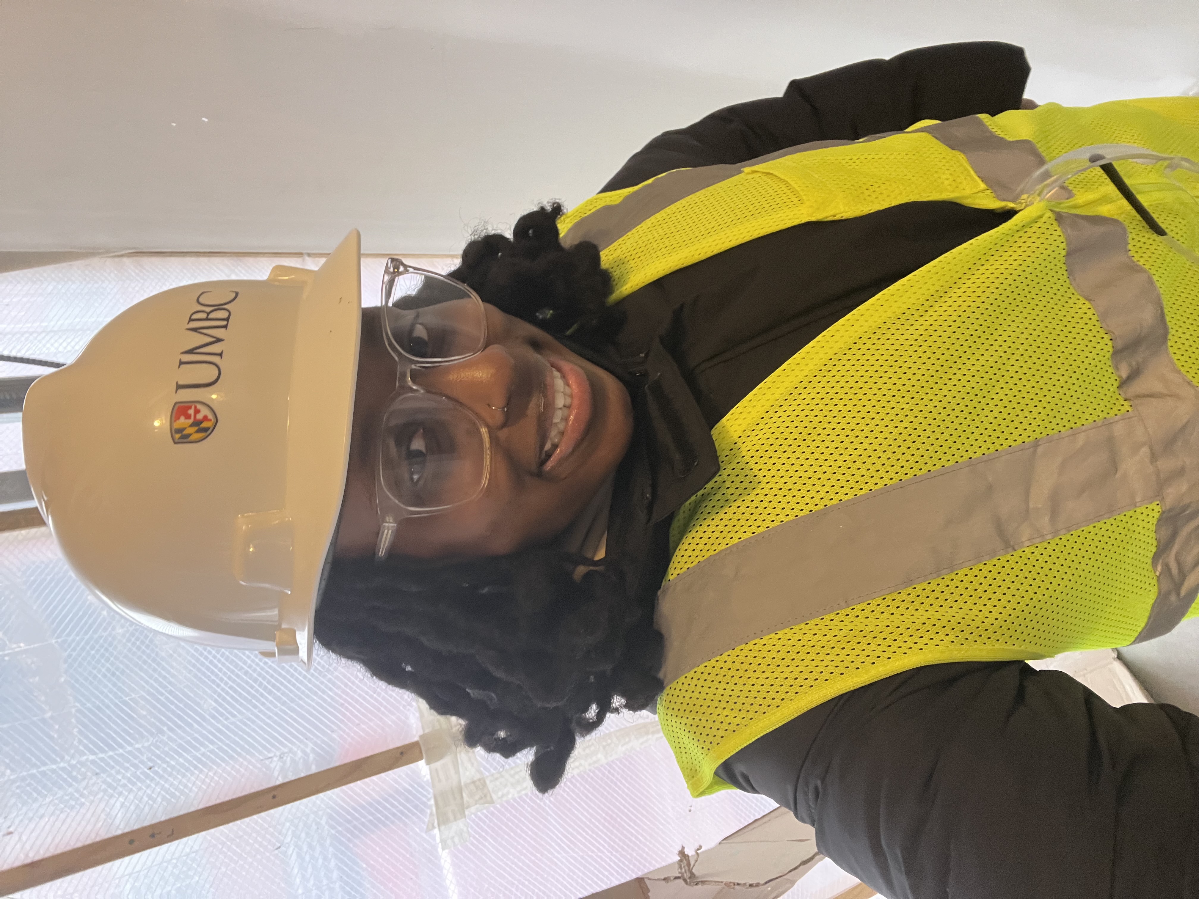 Aissatou in a UMBC white hardhat along with a safety jacket, smiling standing in front of a window with construction outside