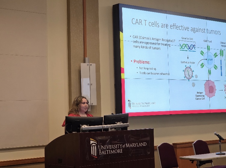 Nicole presenting research while standing behind a black podium with the words university of Maryland Baltimore with research on tv next to her with pictures and the title "CAR T cells are effective against tumors"