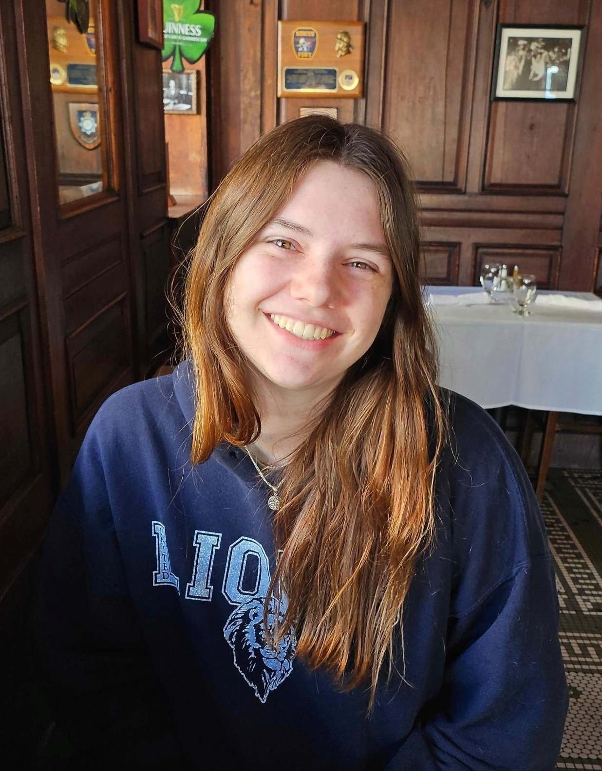 Lucy smiling with a blue sweatshirt with a lion on it sitting in space with brown walls and pictures on them