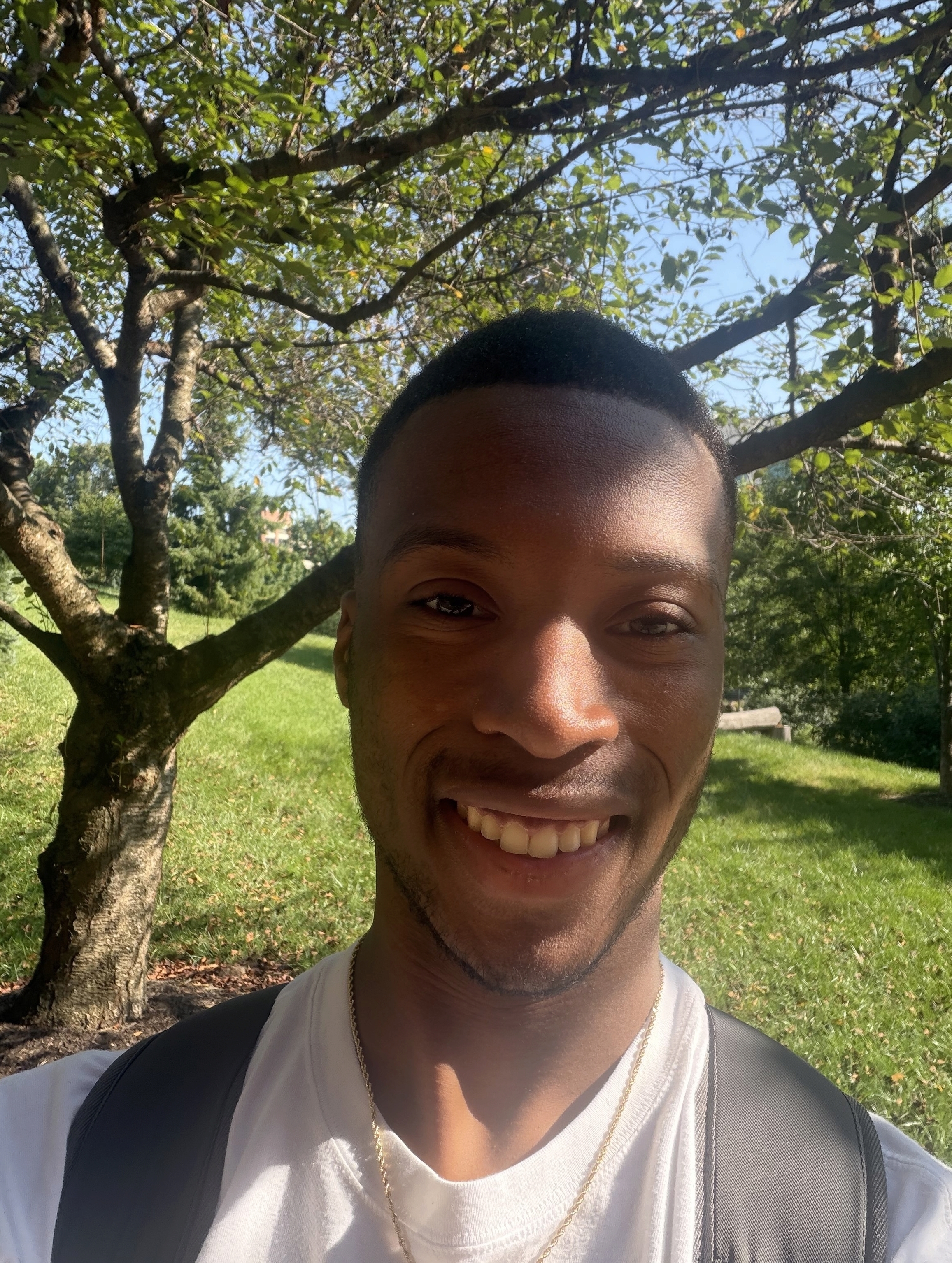 Rhyon is standing in front of a tree and and grassy area smiling in a white t-shirt