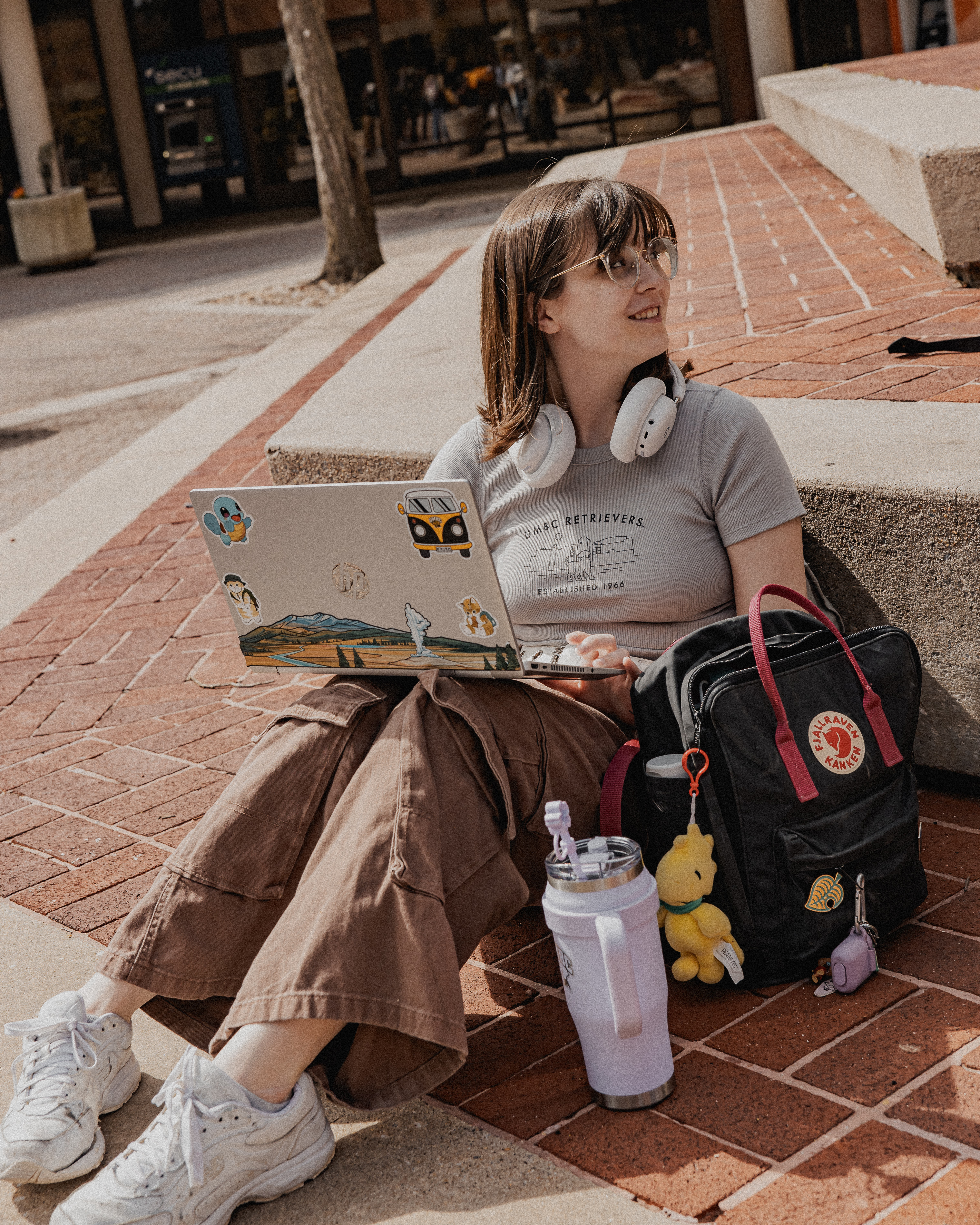 Sofia sitting on the steps outside of UMBC University Center with backpack, water bottle, near her, with laptop on lap and headphones around neck, wearing UMBC Retrievers shirt, looking to the left smiling