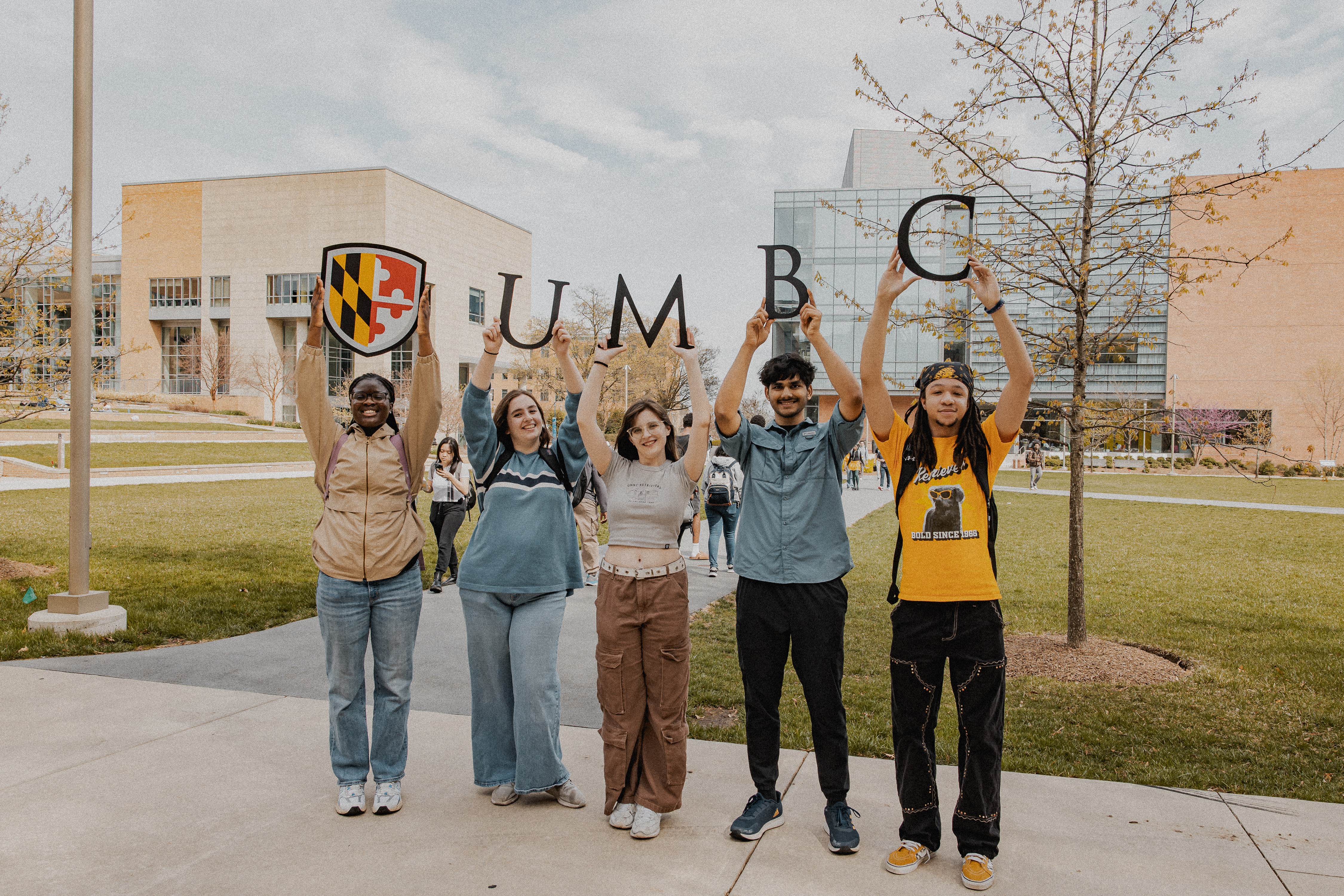 Students standing on pathway hold the UMBC logo and the other students holding up each letter of UMBC