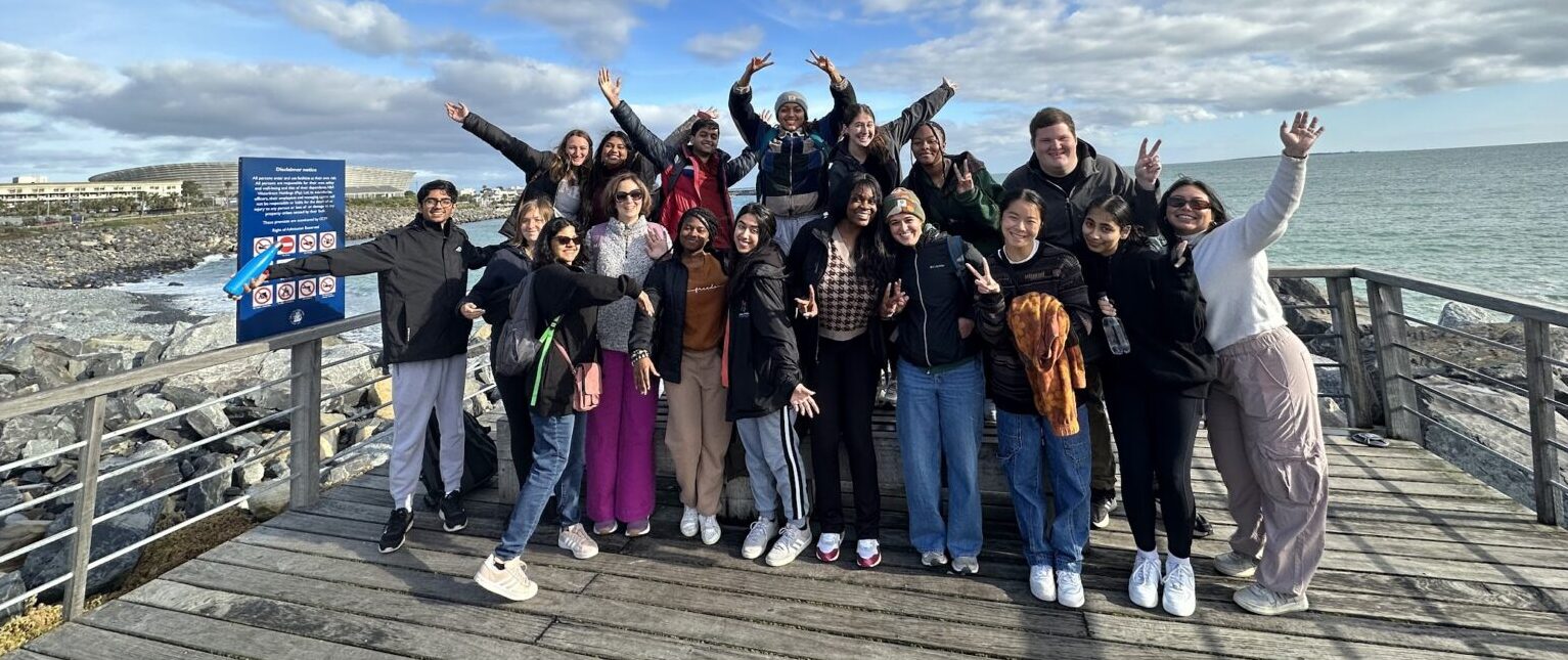 A group of about twenty students waving from a wooden pier with ocean and rocky coast behind them.