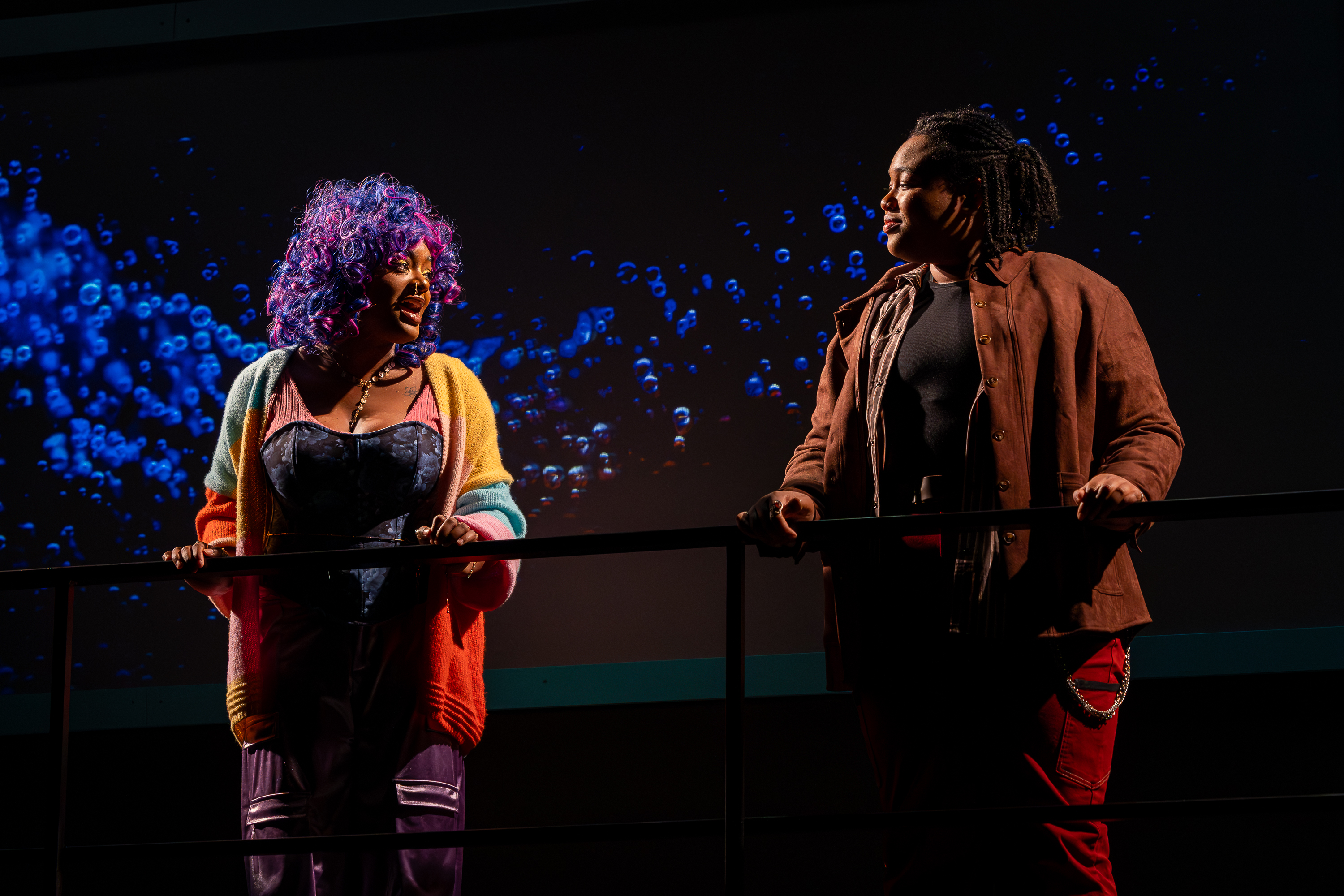 Production photo from User Agreement. Two femme presenting student actors stand behind a railing with a projection of bubbles behind them. They are facing each other, speaking.