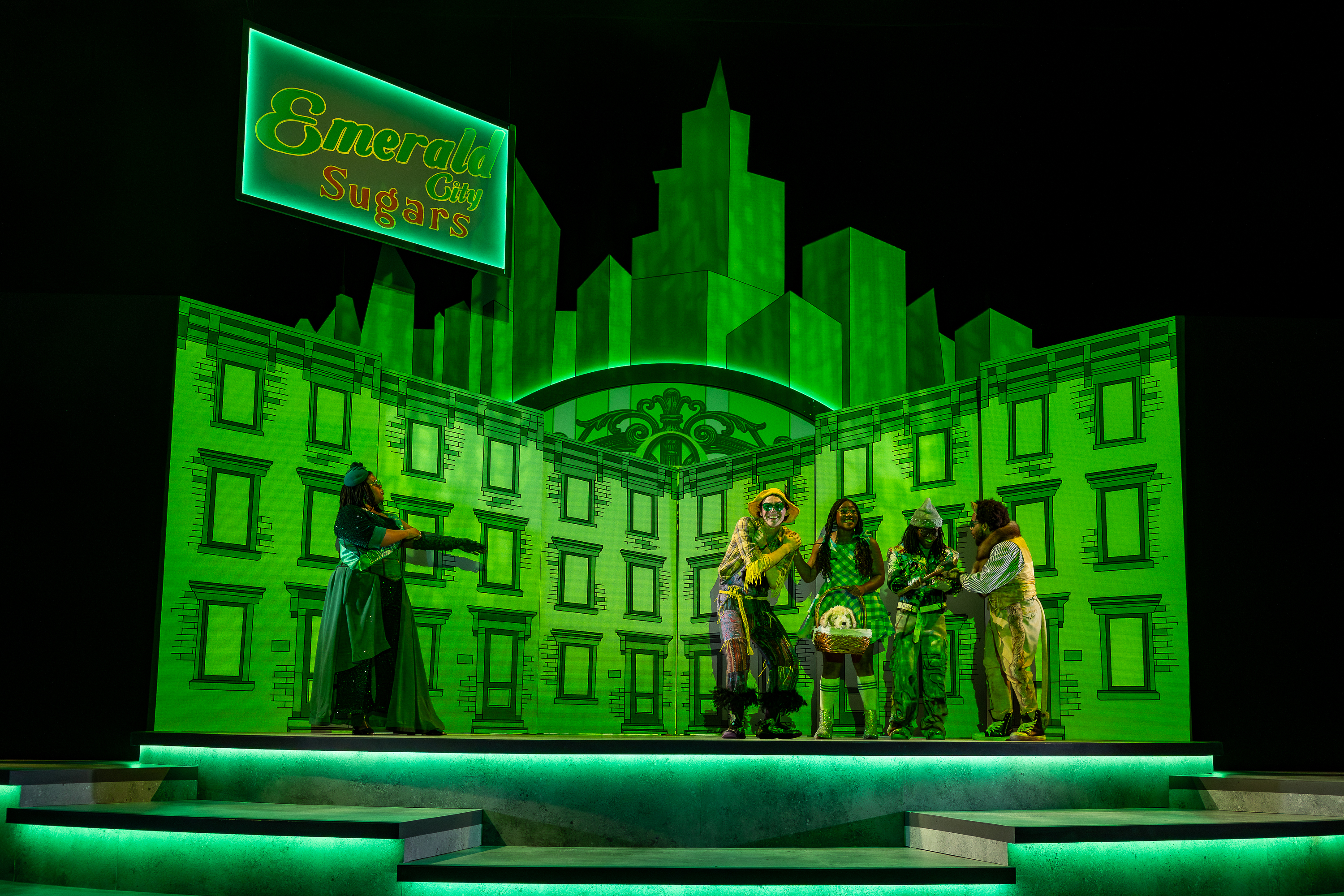 Production Photo from The Wonderful Wizard of Oz. Student actors playing Dorothy, the Scarecrow, the Cowardly Lion, and the Tinman are met in the Emerald City buy the Guardian of the Gates. The city is green.