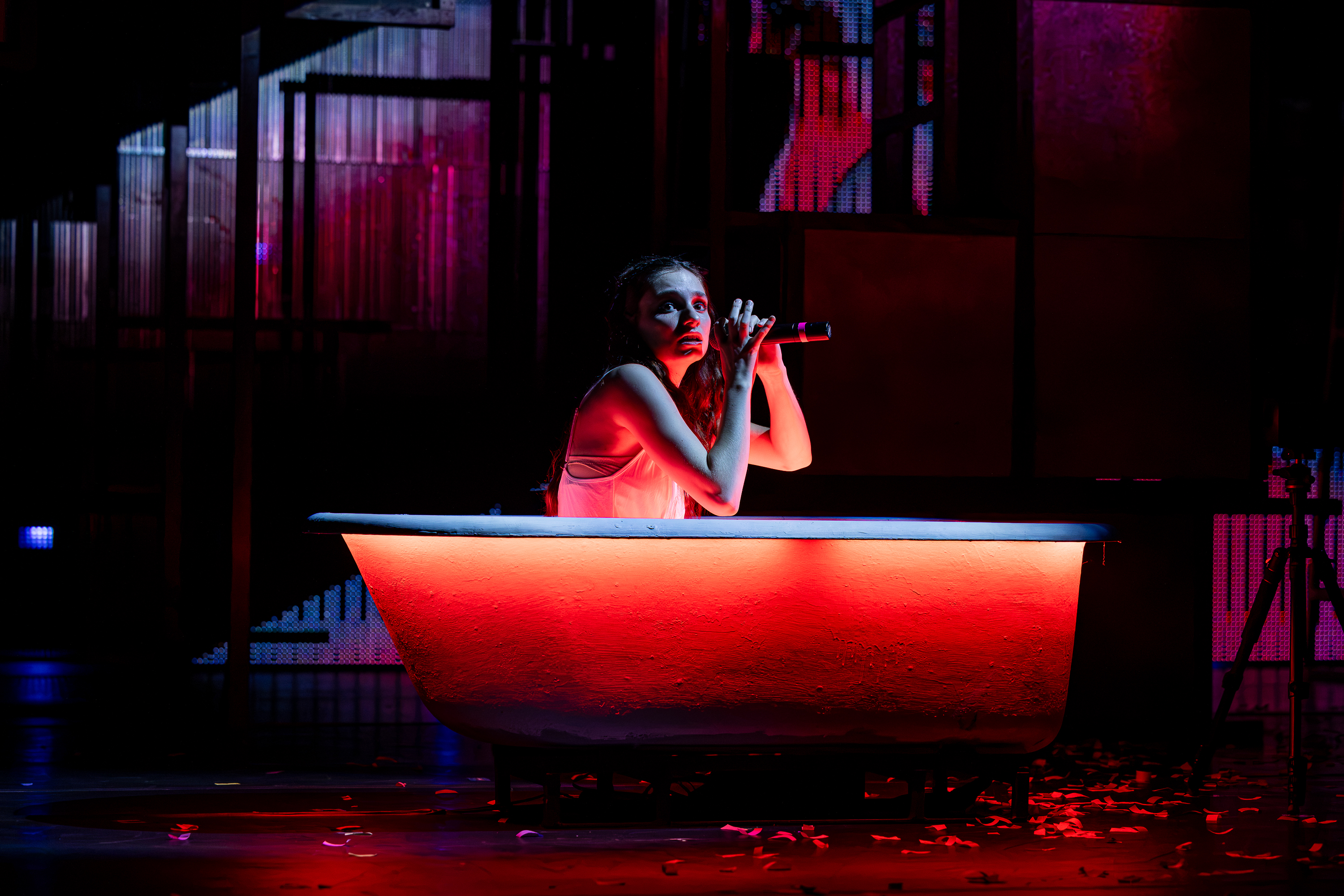 Production photo of a young Lady Macbeth in a bathtub on stage holding a microphone. The stage is illuminated in orange and pink.