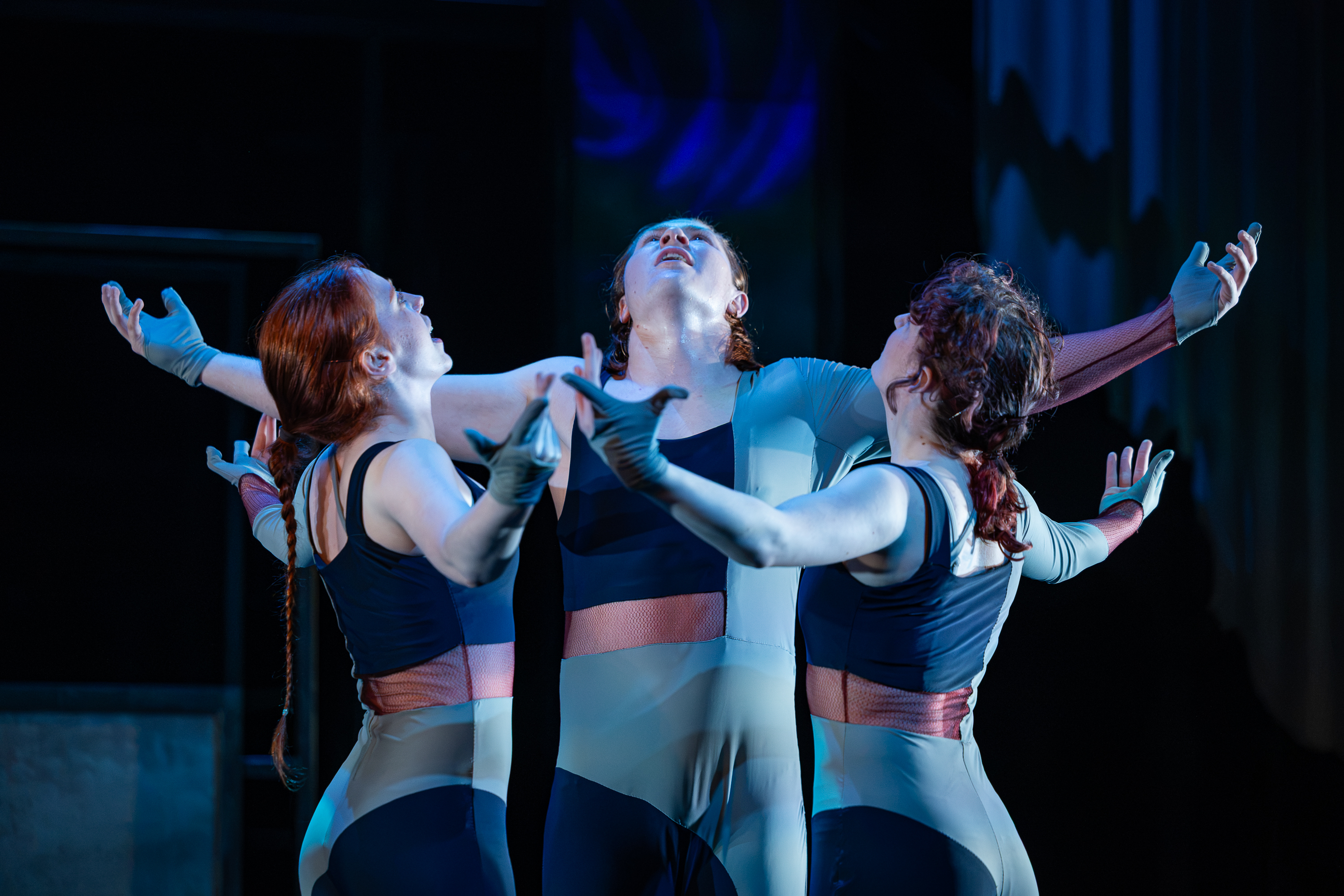 Three student actors dance in character as THEKINDLYONES. They are standing in a circle with their arms spread wide, mouths open, looking at the sky.