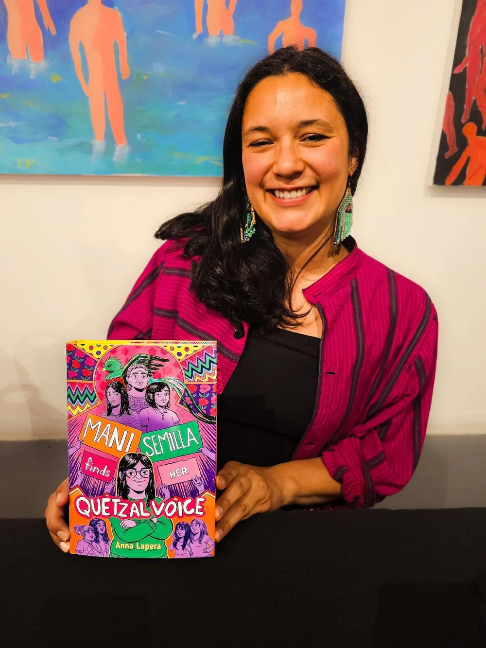 Photo of Anna Lapera in a multi-color jacket holding her book, Mani Semilla Finds Her Quetzal Voice.