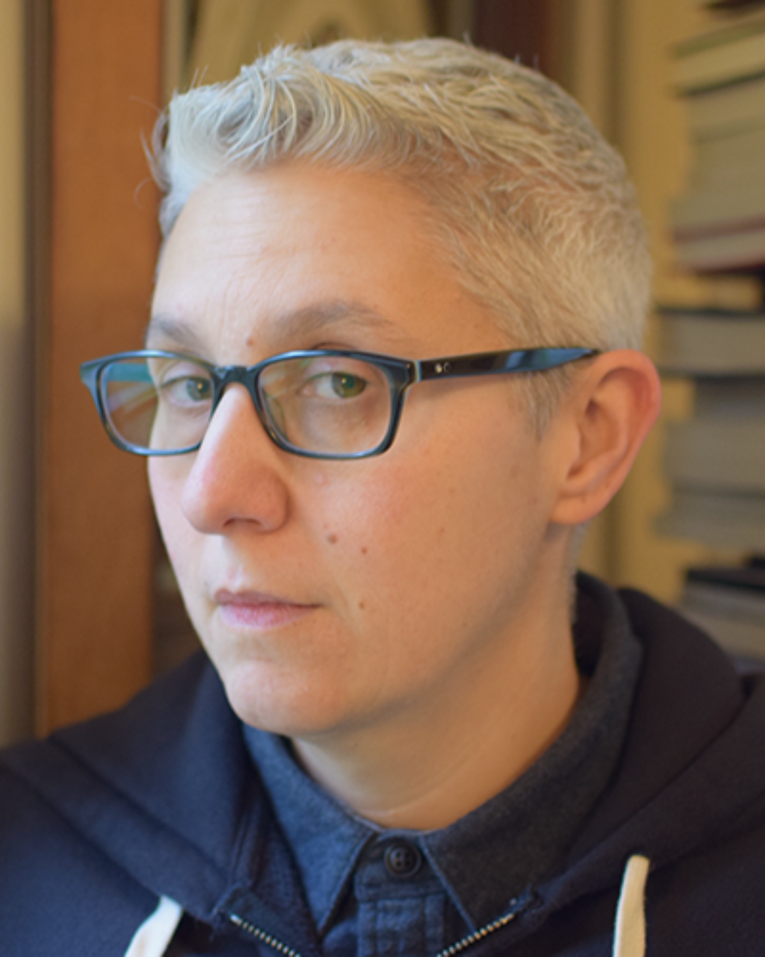 A person with light-skin and short blonde hair is looking forward. They are wearing eyeglasses and a dark colored top.