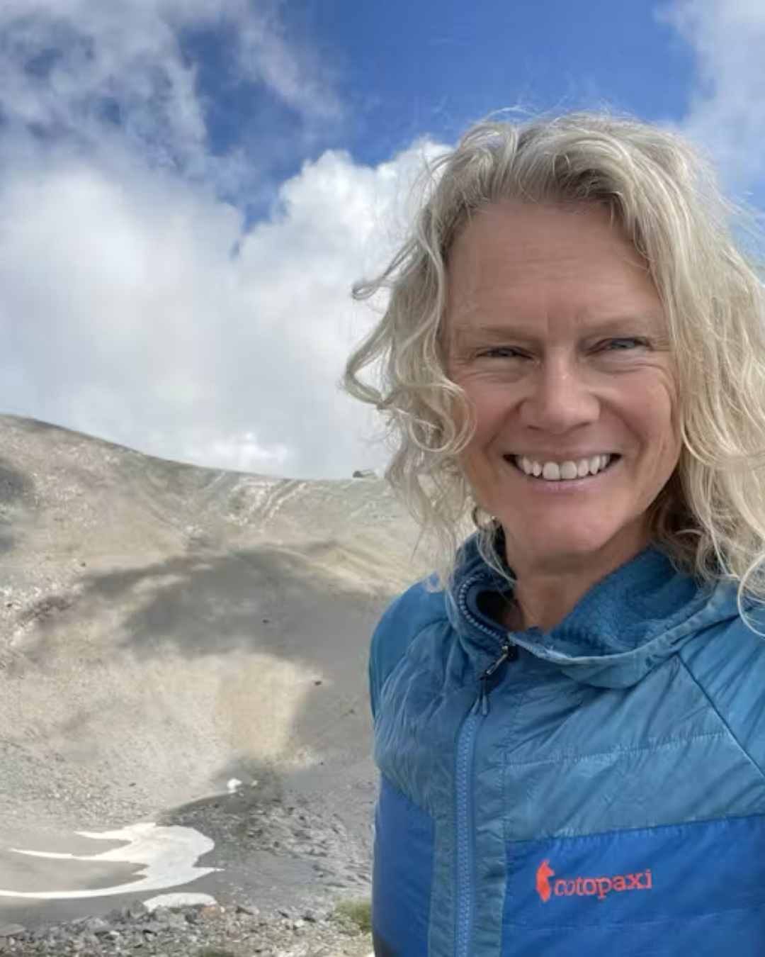 A white woman with blond hair is smiling while standing outside in front of a sand dune.
