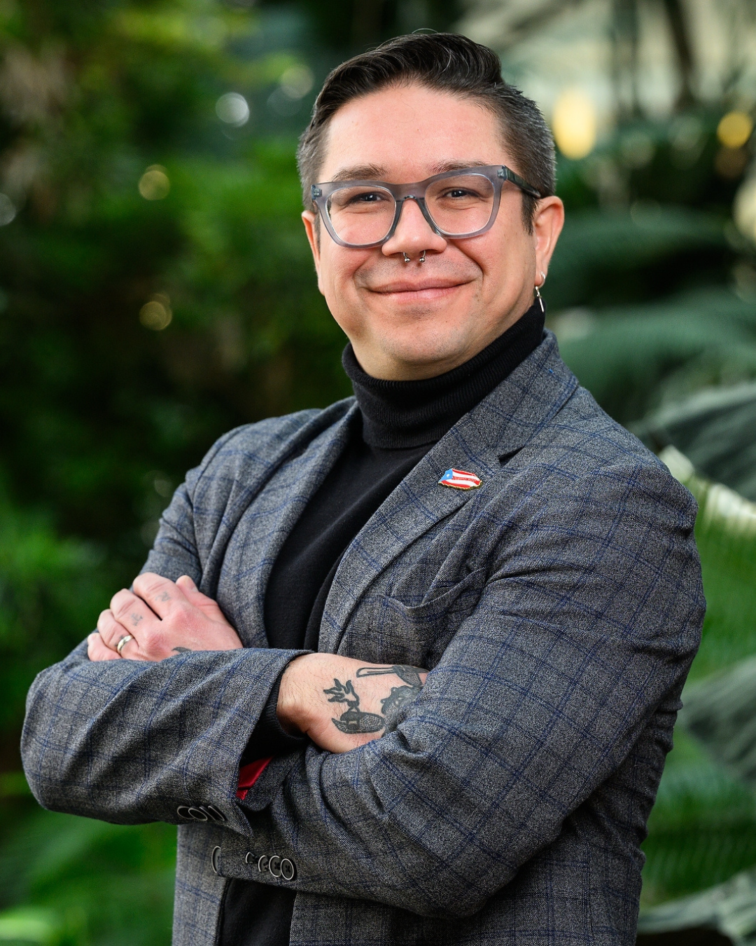 A Puerto Rican man wearing a suit jacket and glasses is standing in front of lush green plants. His arms are crossed and he is smiling.