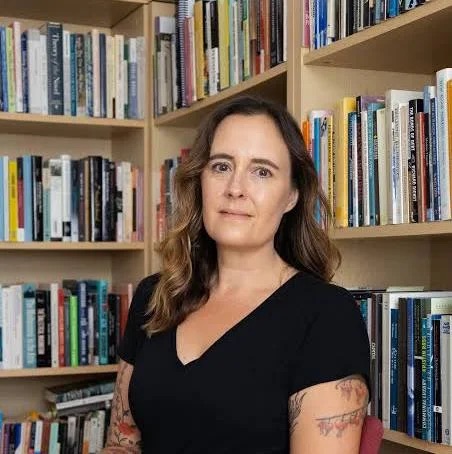 A white woman with long dark hair is seated in front of a bookshelf of books.