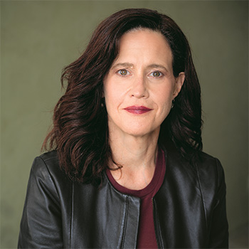 A white woman with reddish hair is looking at the camera. She is wearing a black leather jacket over a red top.