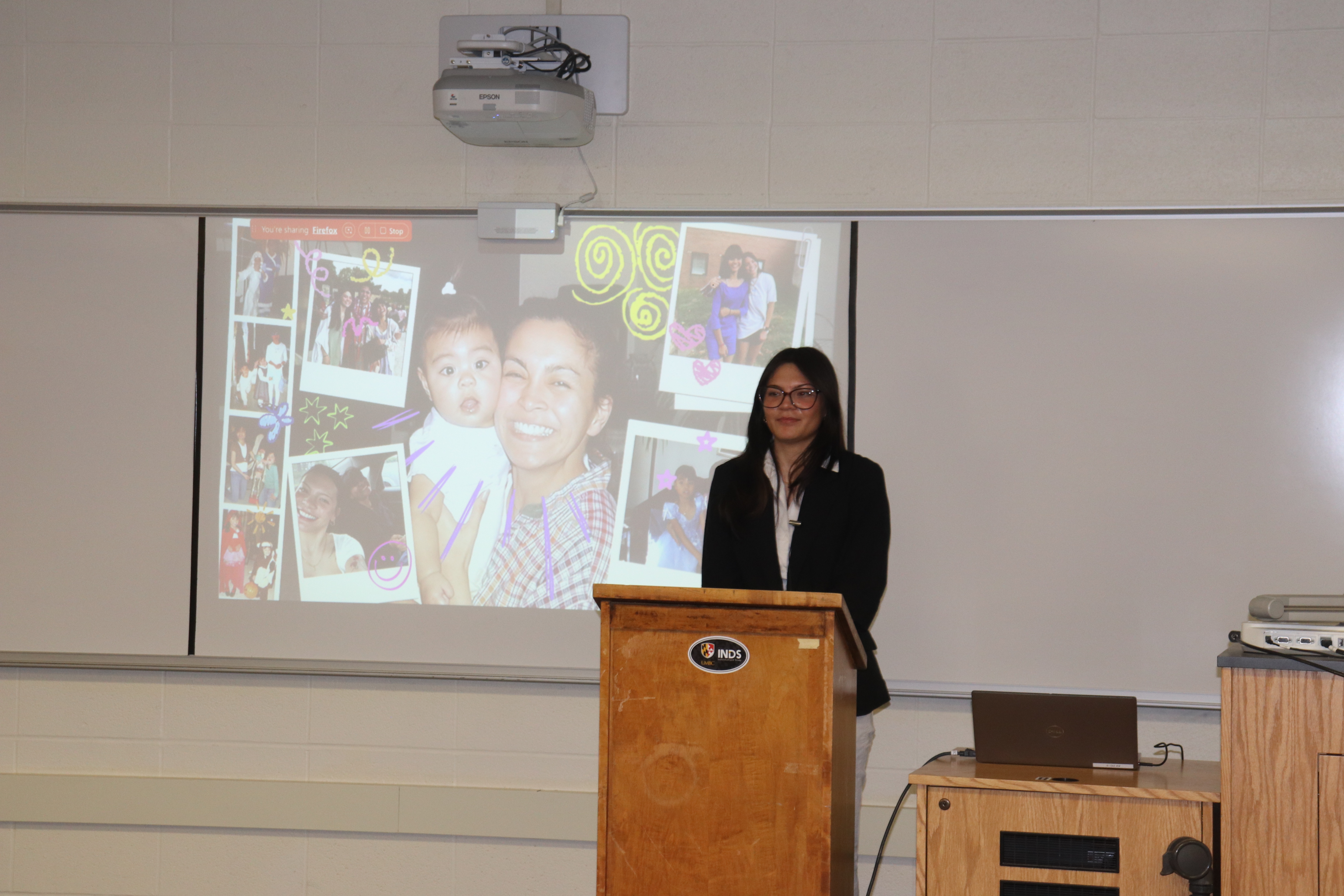 Tristen presenting capstone presentation in black blazer and pants with a white top, with the photos of her sister and mom on the board behind Tristen