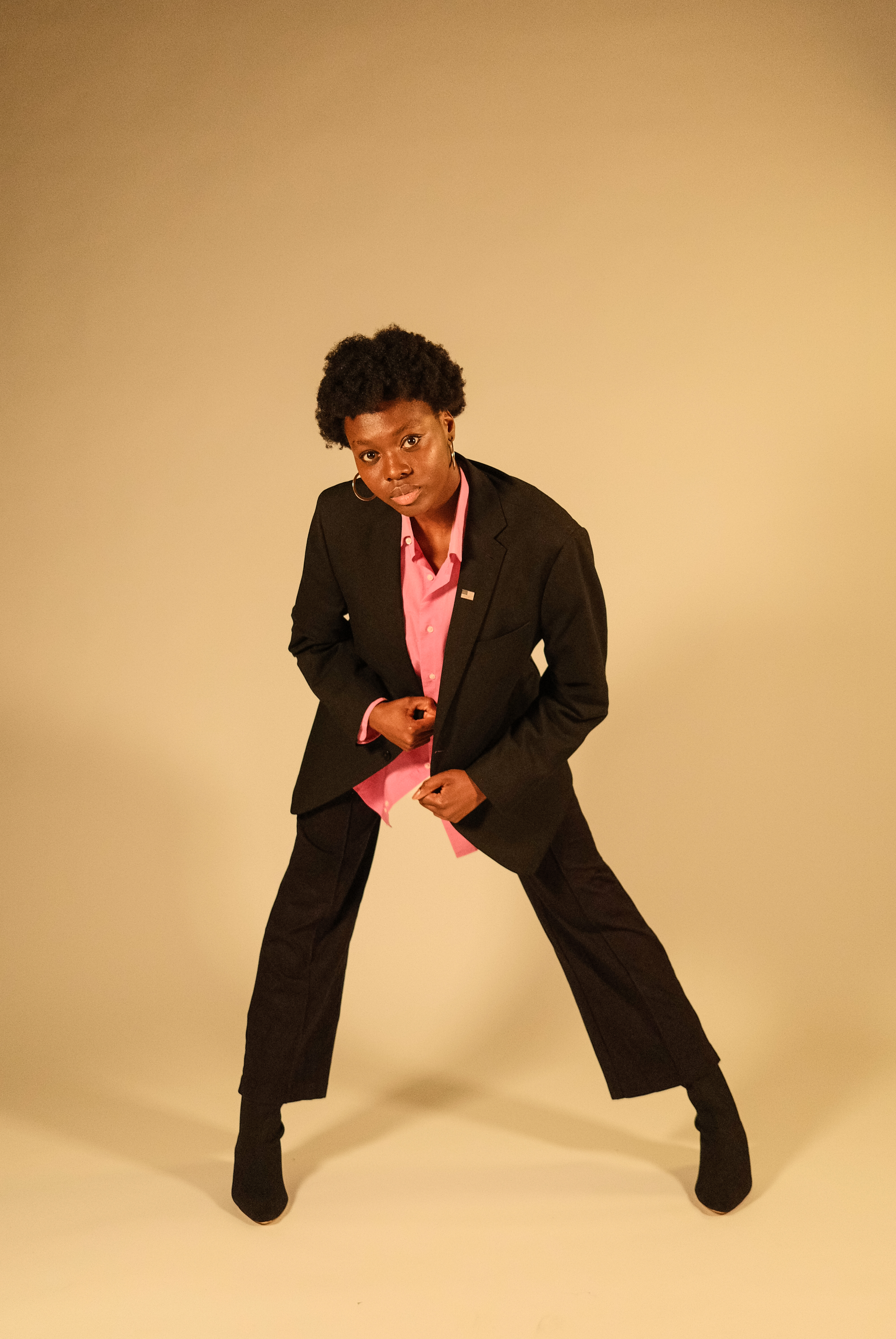 Joanna in a black pant suit with pink button up with a tan background. Joanna is lending towards the camera standing in a dance pose