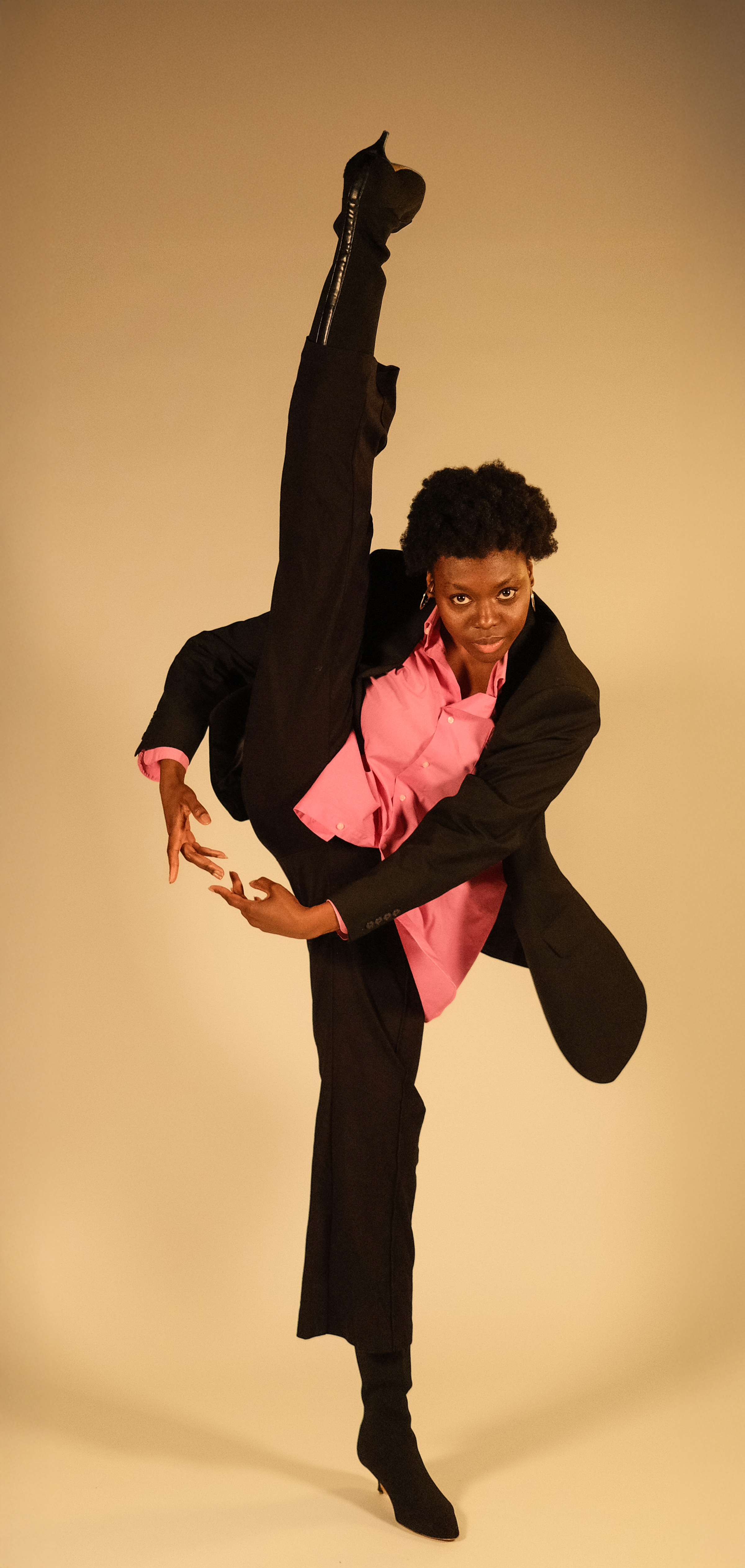 Joanna in a black pant suit with pink button up standing on one leg with the other leg straight in the air showing a black shoe with small heel, with hands between legs in a dance pose