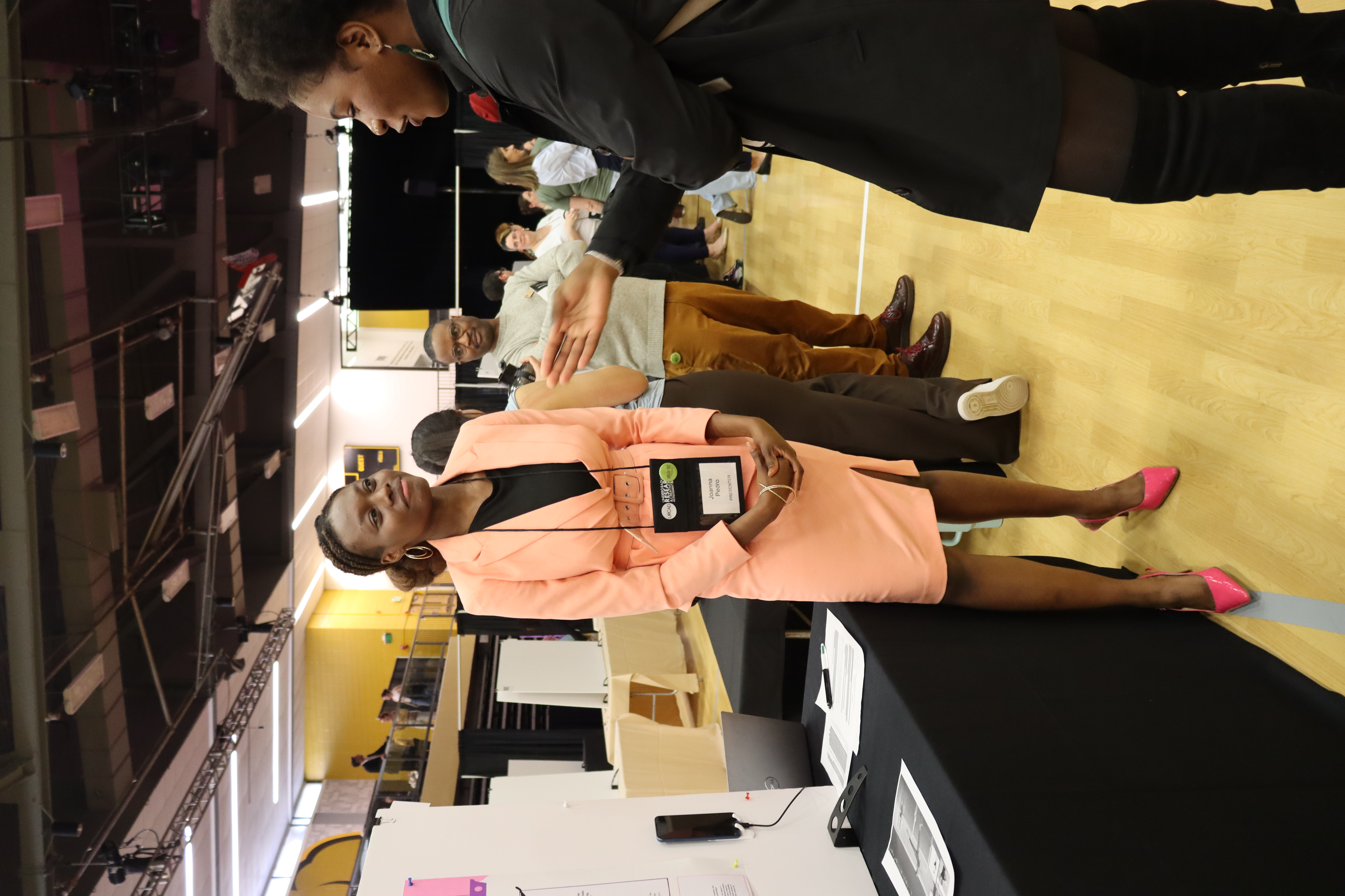 Joanna in a peach business suit standing next to URCAD poster and research, looking at student who is looking at Joanna's URCAD project