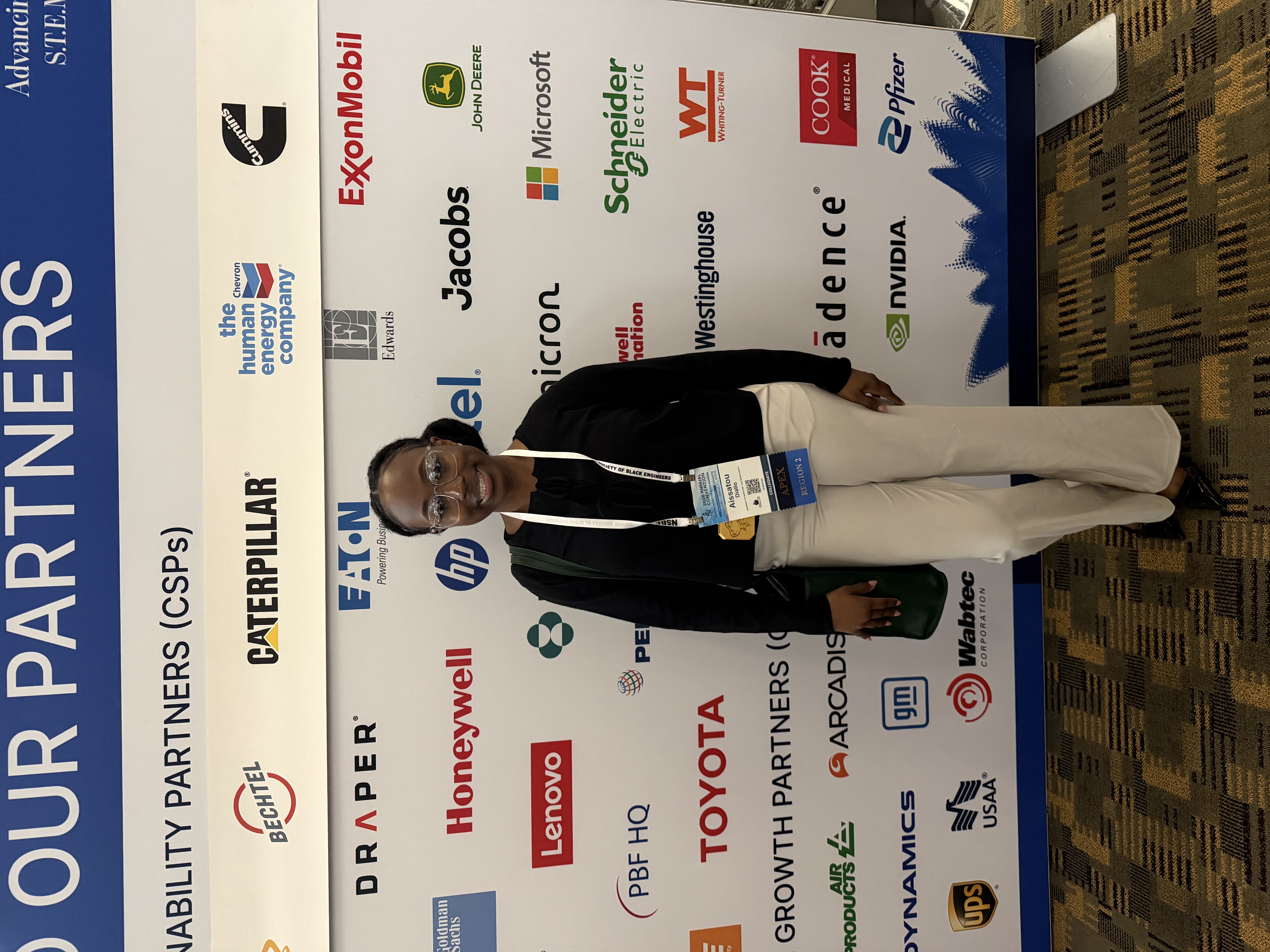 Aissatou in a black top and white pants smiling in front of a background with names of multiple partners to the NSBE conference