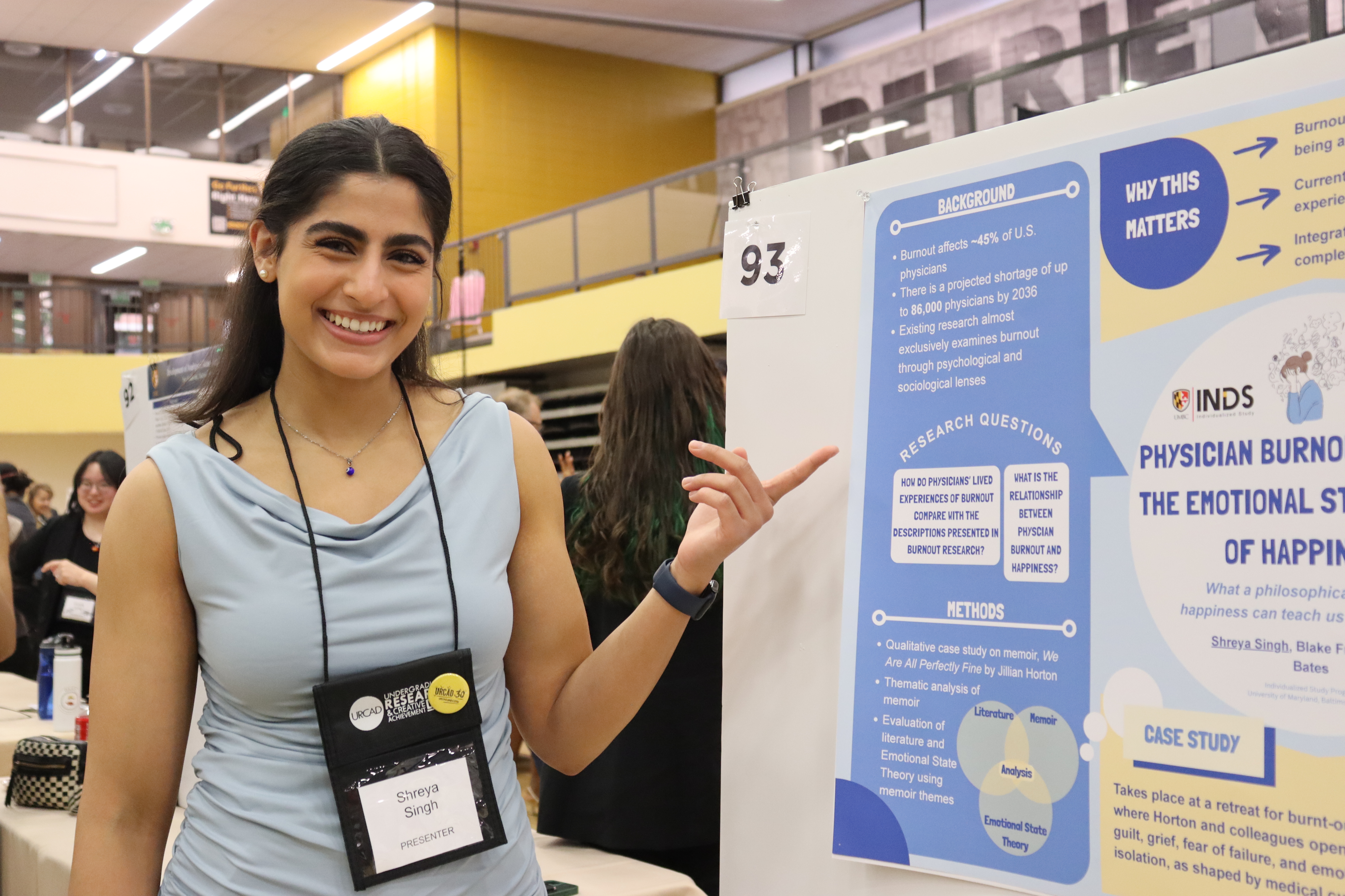 Shreya smiling in a blue top next to URCAD poster
