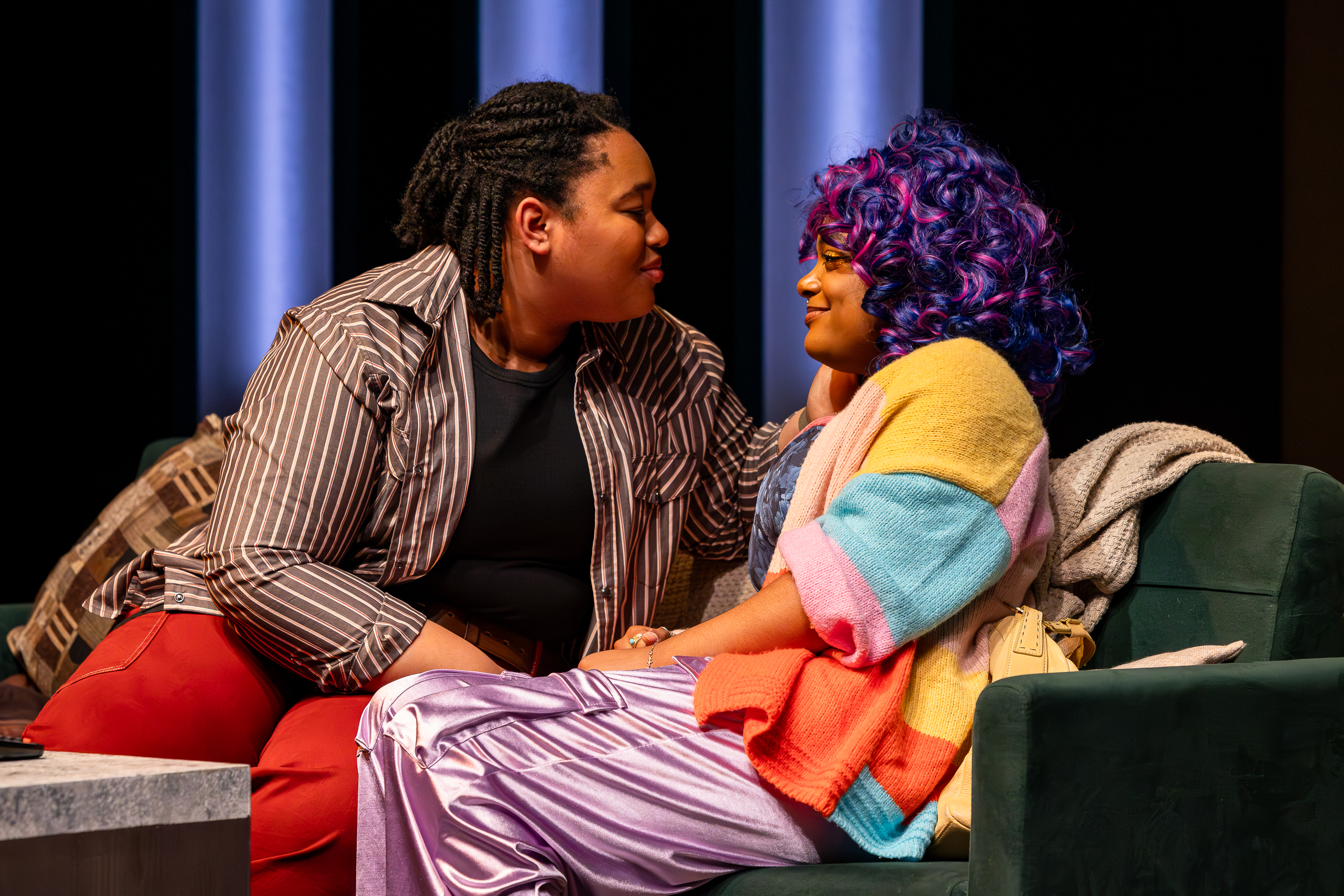 Production photo from User Agreement. Two Black femme presenting student actors sit on a couch and look lovingly into each other's eyes.
