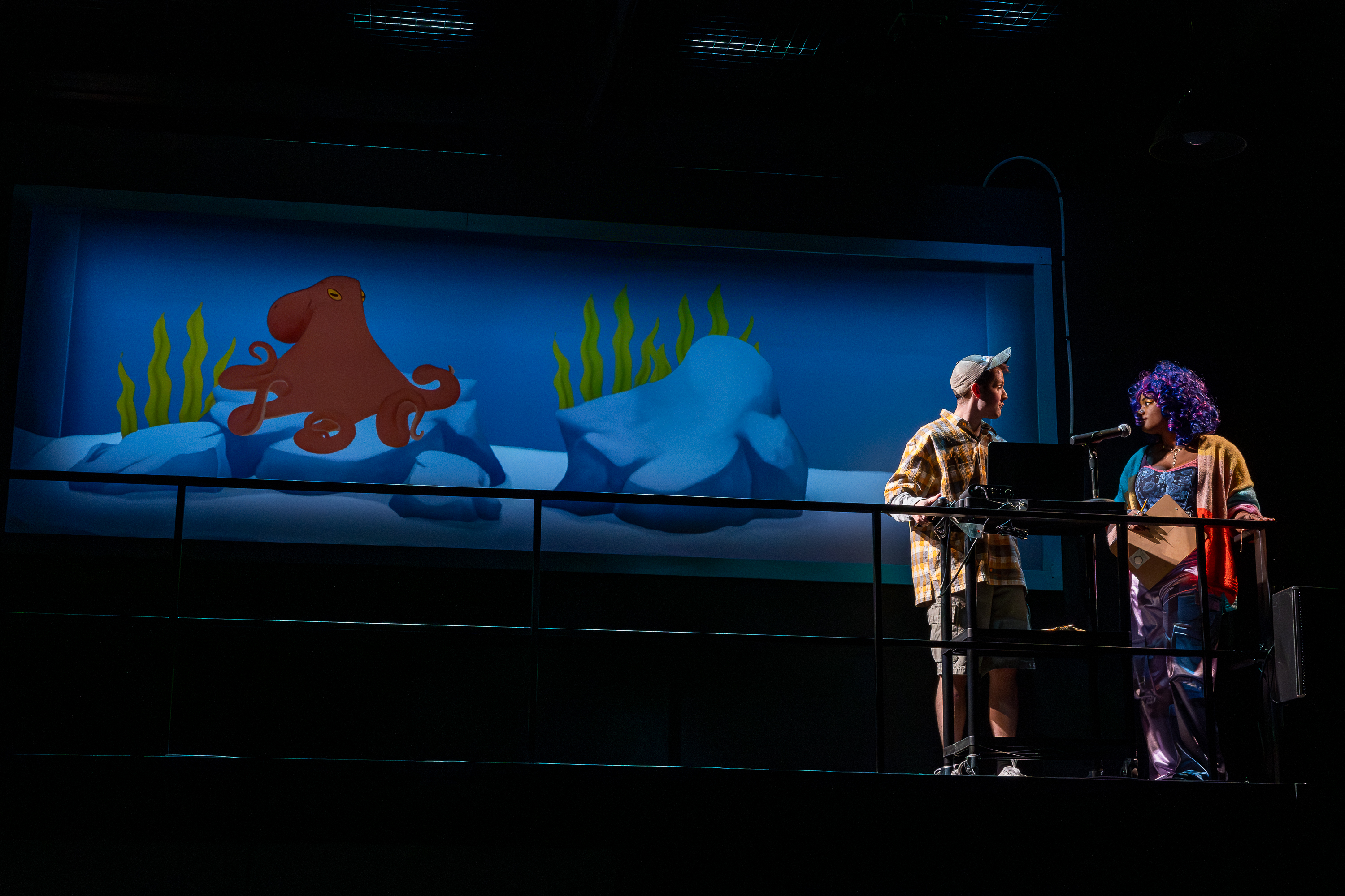 Production photo from User Agreement. Two characters try to speak with an animated octopus that is projected to the side of them. The octopus is red in an aquarium tank.