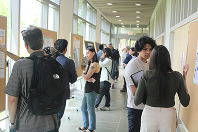 Students and faculty standing in a bright hallway, looking at academic posters pinned to large corkboards during a competition.